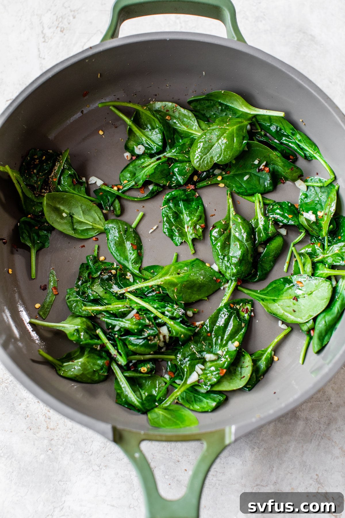 A close-up of wilted spinach, minced garlic, and red pepper flakes sizzling in olive oil within a stainless steel skillet.