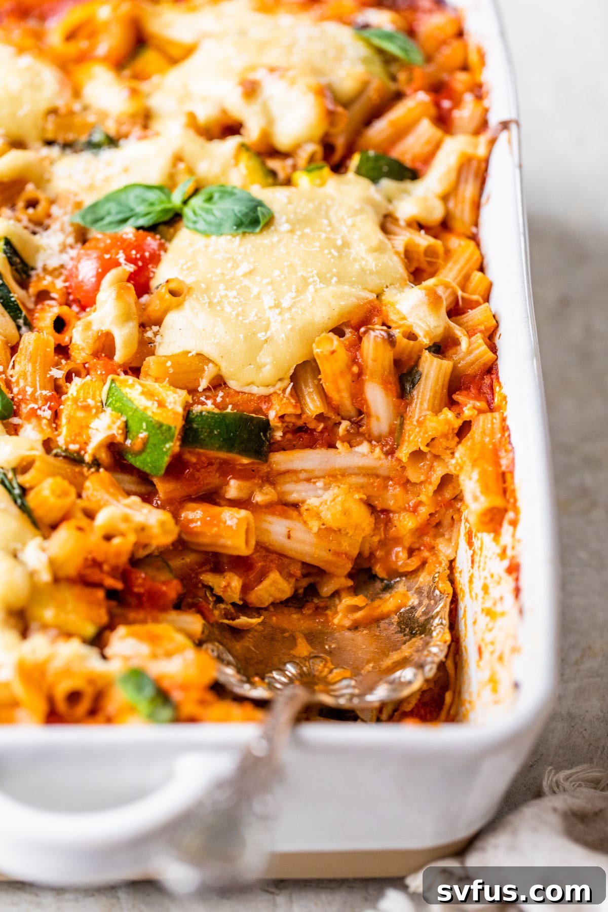 A freshly baked vegan pasta casserole in a ceramic dish, with a serving spoon resting in it, showcasing the perfectly cooked pasta and golden cashew cheese topping.