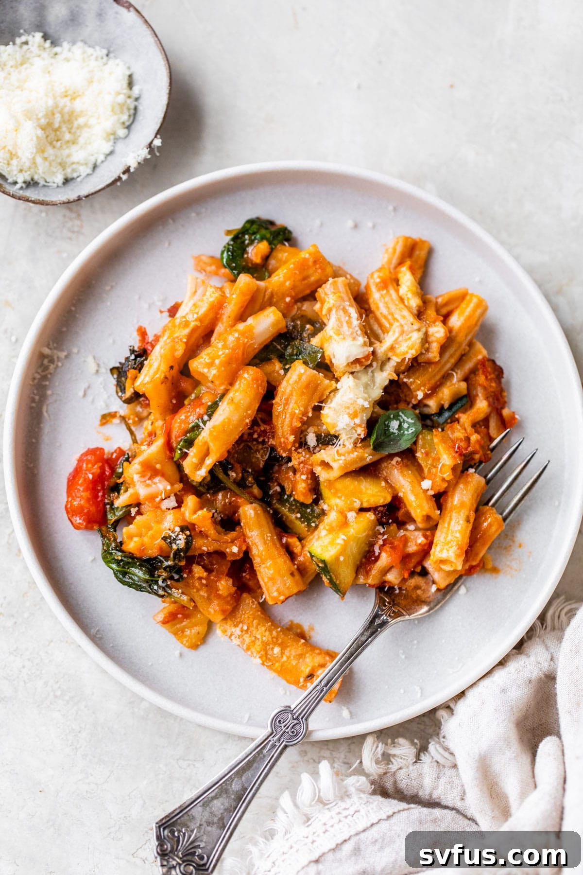 A close-up shot of a single serving of vegan pasta bake on a white plate, showing the rich pasta, zucchini, and tomatoes coated in sauce and topped with creamy cashew cheese.