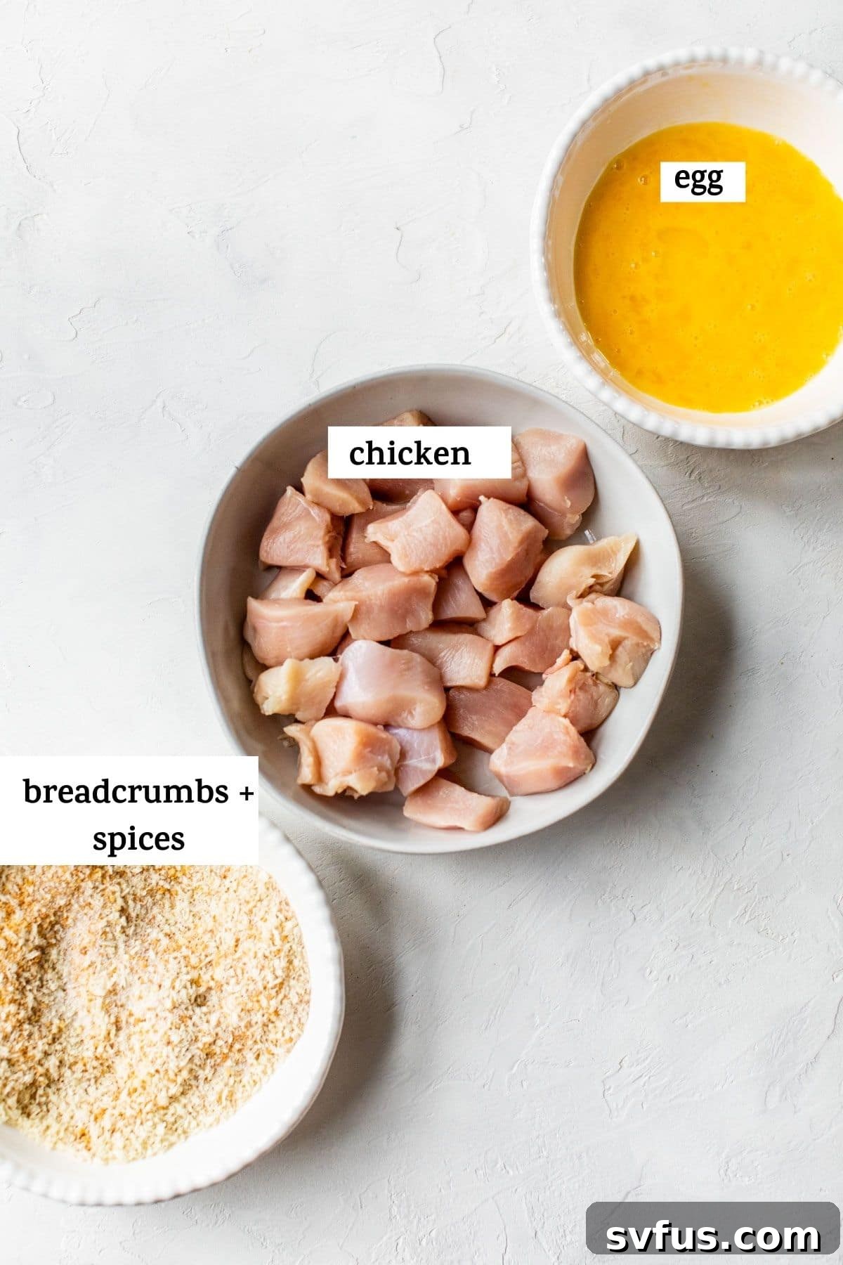 Ingredients for homemade Air Fryer Chicken Nuggets: bowls of breadcrumbs, raw chicken breast pieces, and whisked eggs ready for preparation