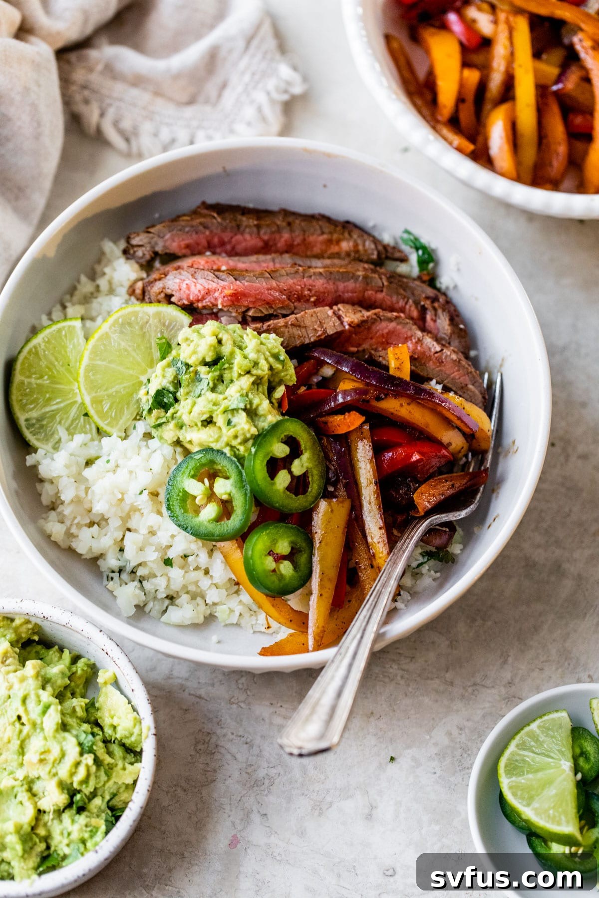 A beautifully arranged steak fajita bowl featuring cilantro lime cauliflower rice as the base, topped with perfectly grilled flank steak, vibrant bell peppers, and a creamy dollop of guacamole, presented against a rustic background.