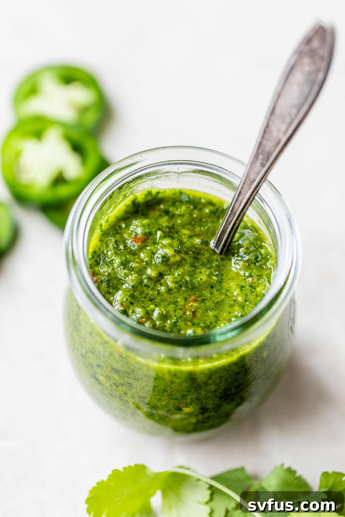 A elegant glass jar filled with spicy green Zhoug sauce, with a small spoon emerging from its depths, set against a simple background.