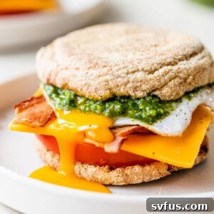 english muffin on a plate topped with tomato, sliced cheese, fried egg, and bacon