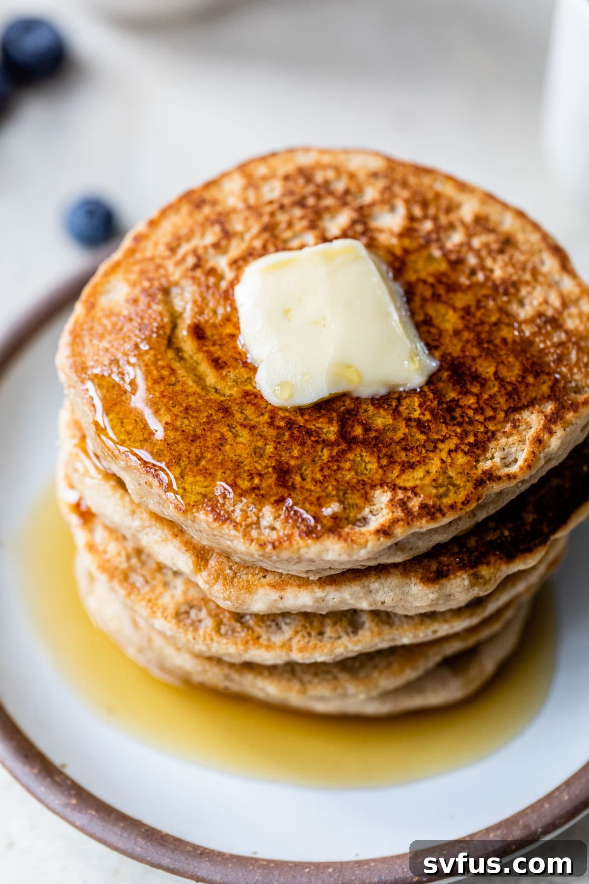 A stack of fluffy oat flour pancakes topped with a pat of butter and a generous pour of golden maple syrup, ready to be enjoyed.