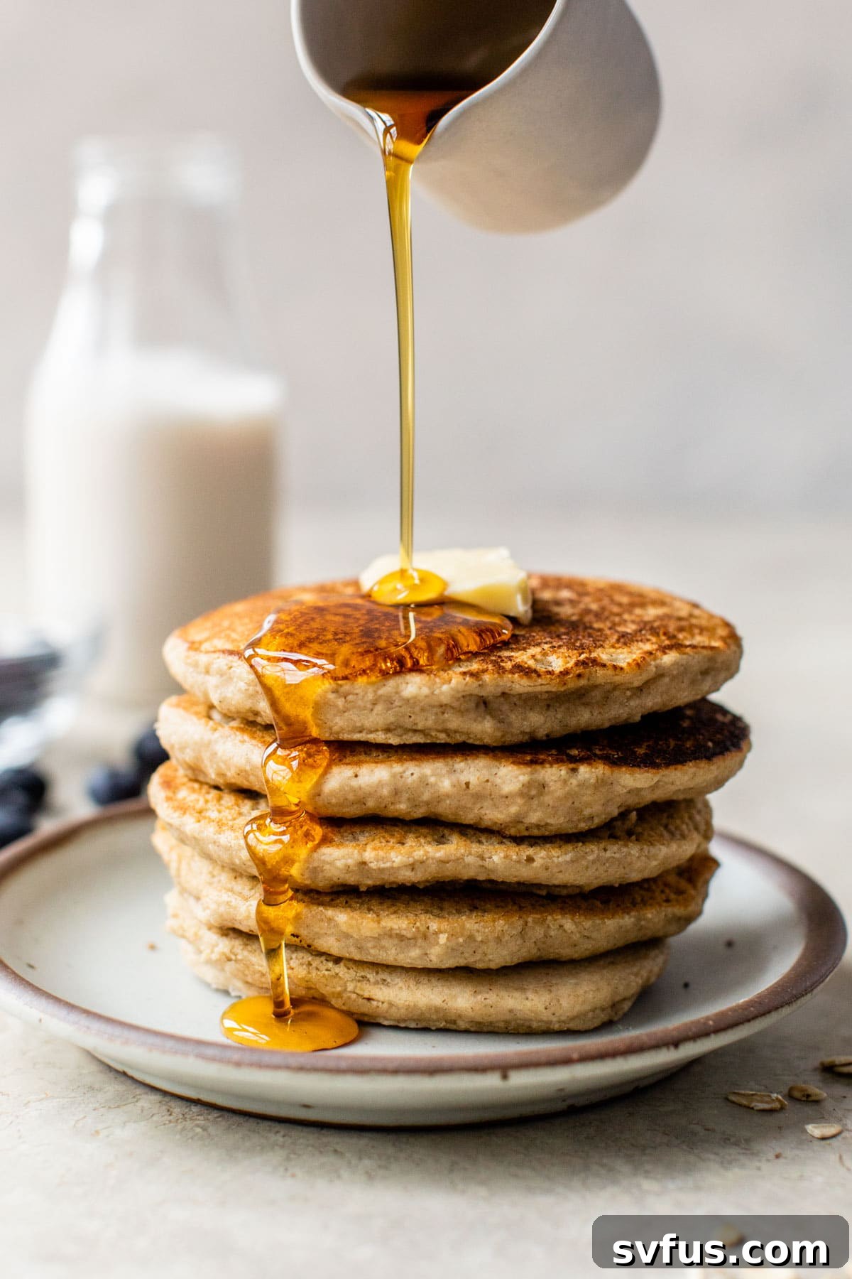 Someone is gently pouring golden maple syrup over a freshly made stack of fluffy oat flour pancakes, highlighting their soft texture and delicious appeal.