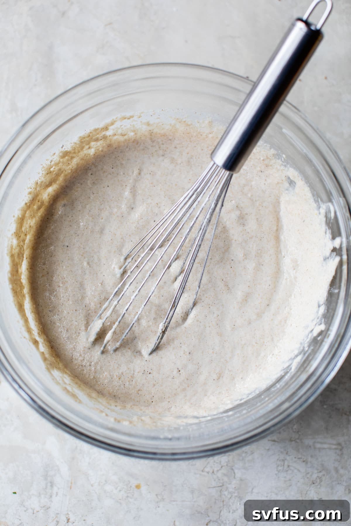 Close-up of thick, light beige oat flour pancake batter in a clear glass mixing bowl, showcasing its smooth consistency before cooking.