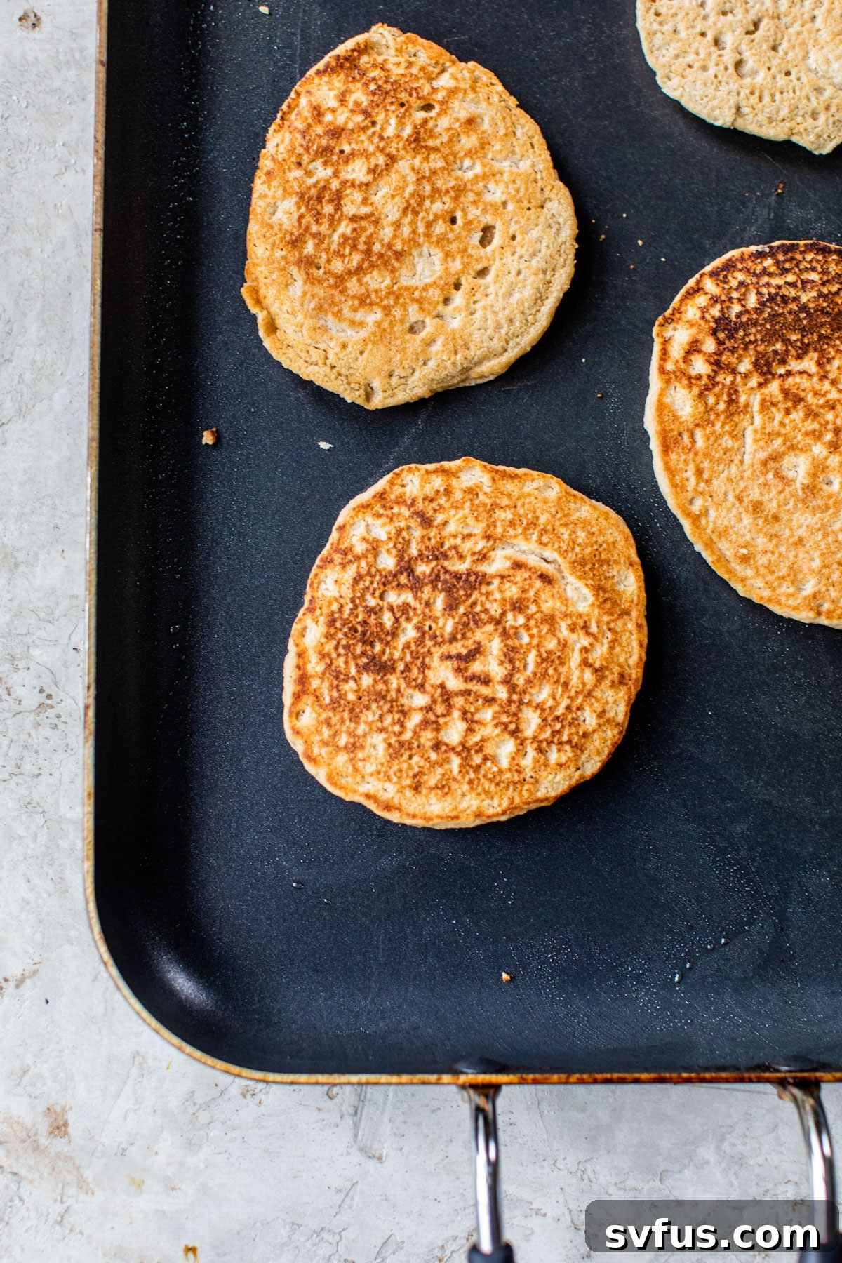 Several golden-brown oat flour pancakes sizzling on a heated griddle, some with small bubbles indicating they are ready to flip.
