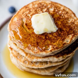 A close-up of a perfectly stacked pile of fluffy oat flour pancakes, garnished with a dollop of butter and drizzled generously with maple syrup.