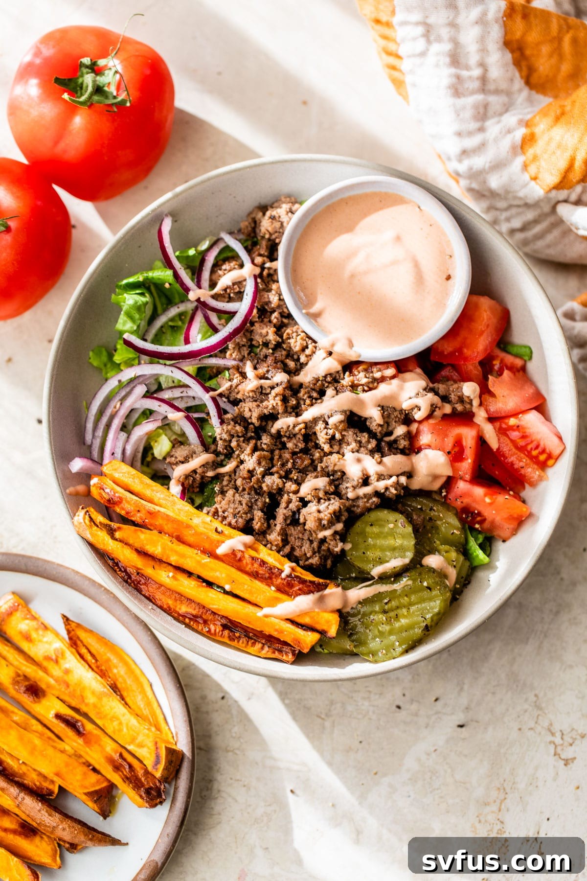 A close-up shot of a prepared burger bowl, showcasing the ground beef, sweet potato fries, sliced tomato, red onion, and dill pickles, inviting a delicious meal.