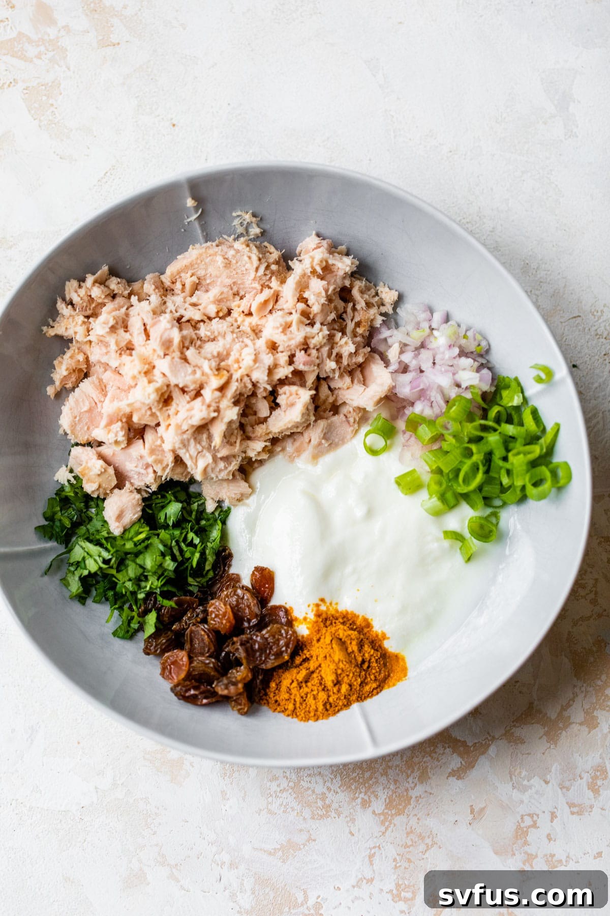 Close-up of a bowl containing drained tuna, creamy Greek yogurt, vibrant cilantro, minced shallot, green onion, and golden raisins, ready to be mixed into a curried tuna salad.