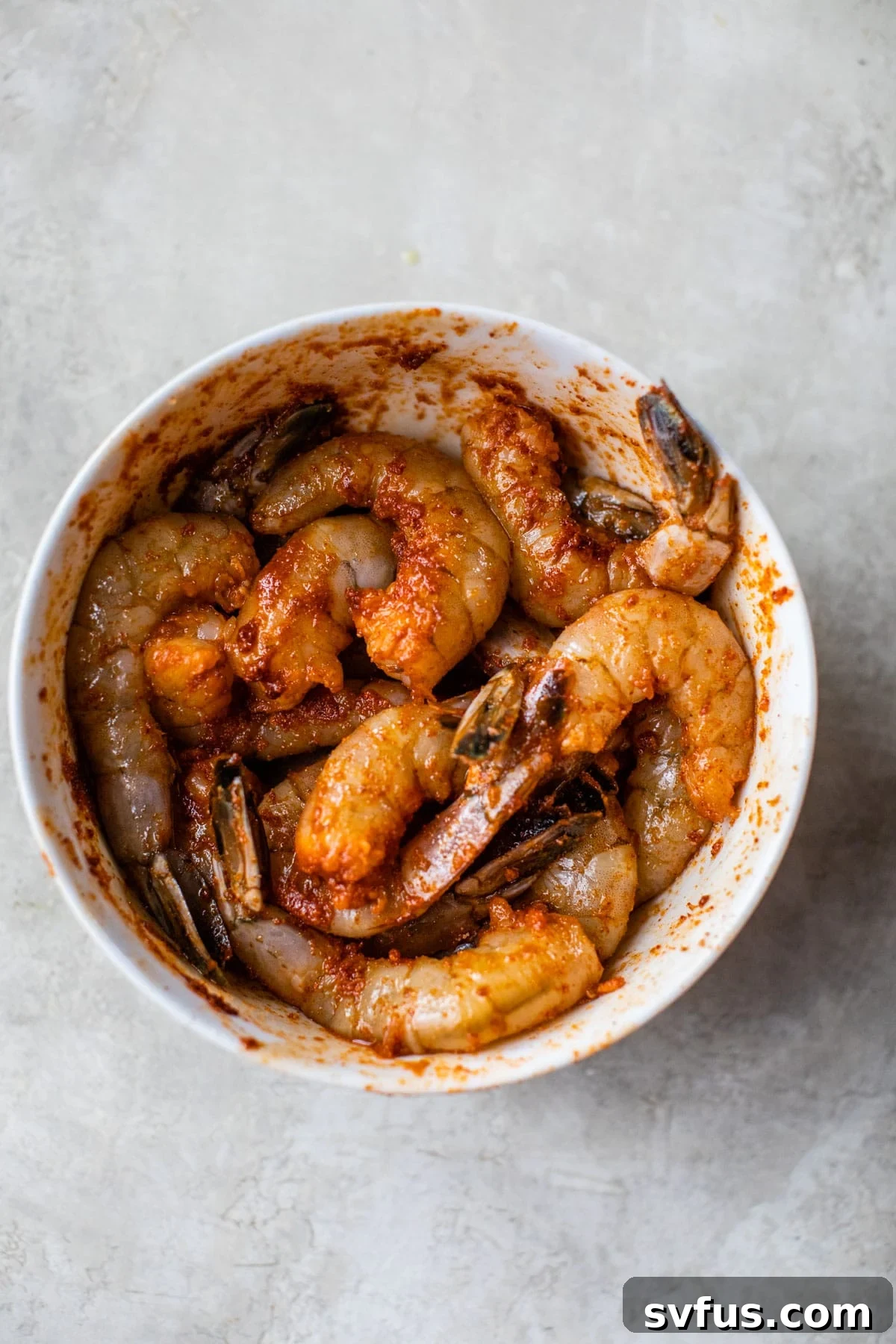 Raw shrimp in a bowl, thoroughly coated with olive oil and a savory spice blend, ready for air frying.
