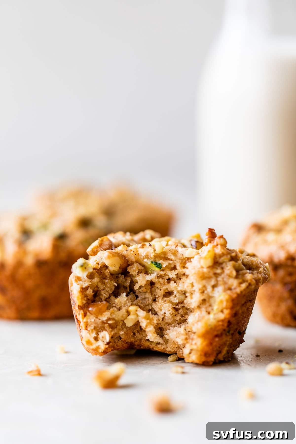 A perfectly baked vegan zucchini muffin rests on a rustic wooden table, with a bite taken out to reveal its tender, moist interior and visible specks of zucchini and walnuts.