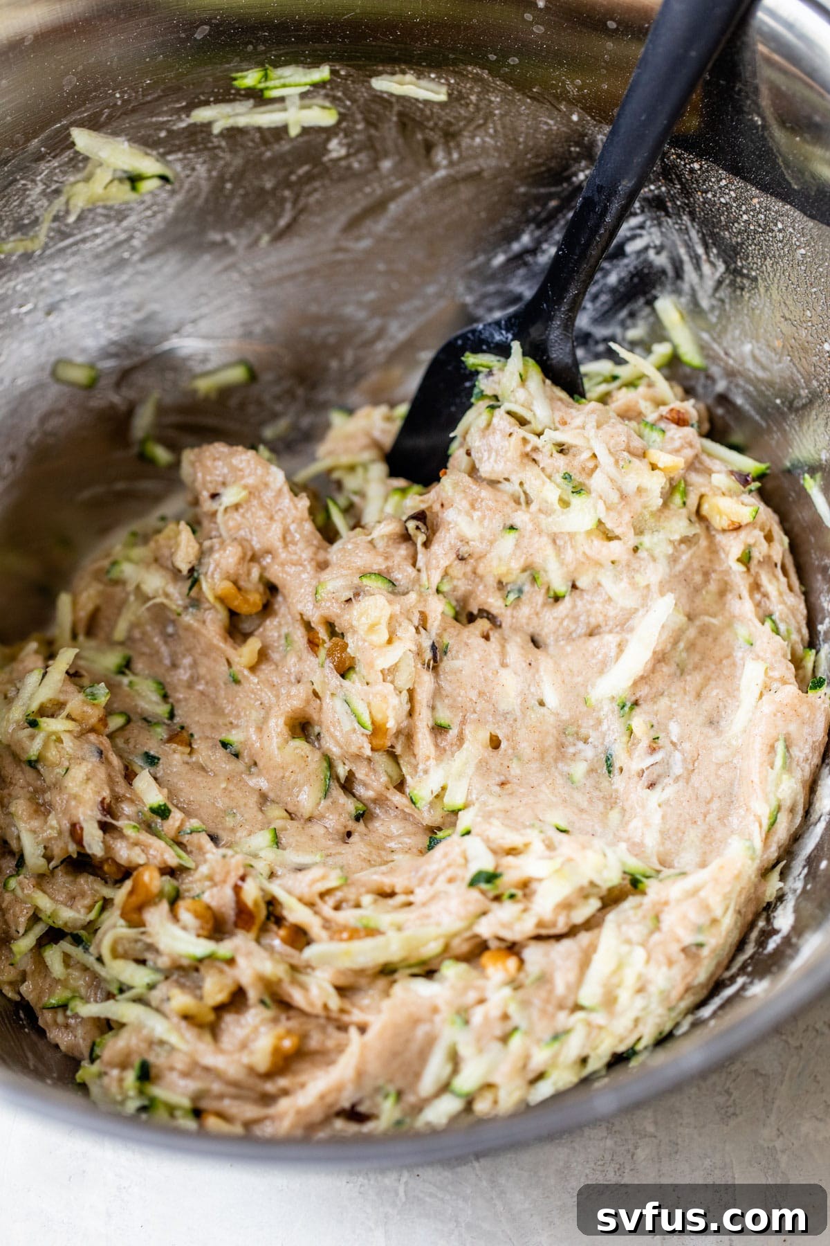 A close-up shot of rich, fluffy vegan zucchini muffin batter in a large mixing bowl, speckled with green zucchini strands and chopped walnuts, ready for baking.