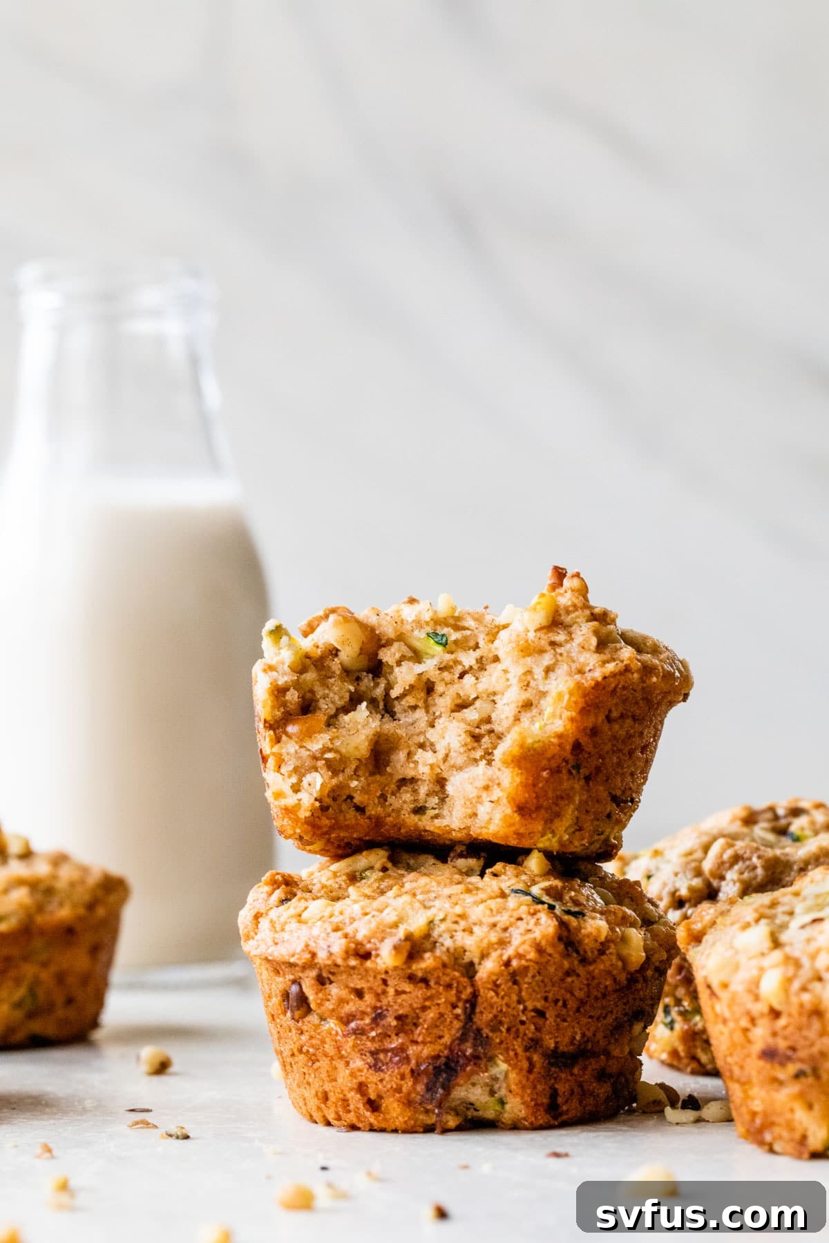 Two soft, golden-brown vegan zucchini muffins are stacked elegantly beside a tall glass of refreshing dairy-free milk, inviting you to enjoy a perfect snack or breakfast.