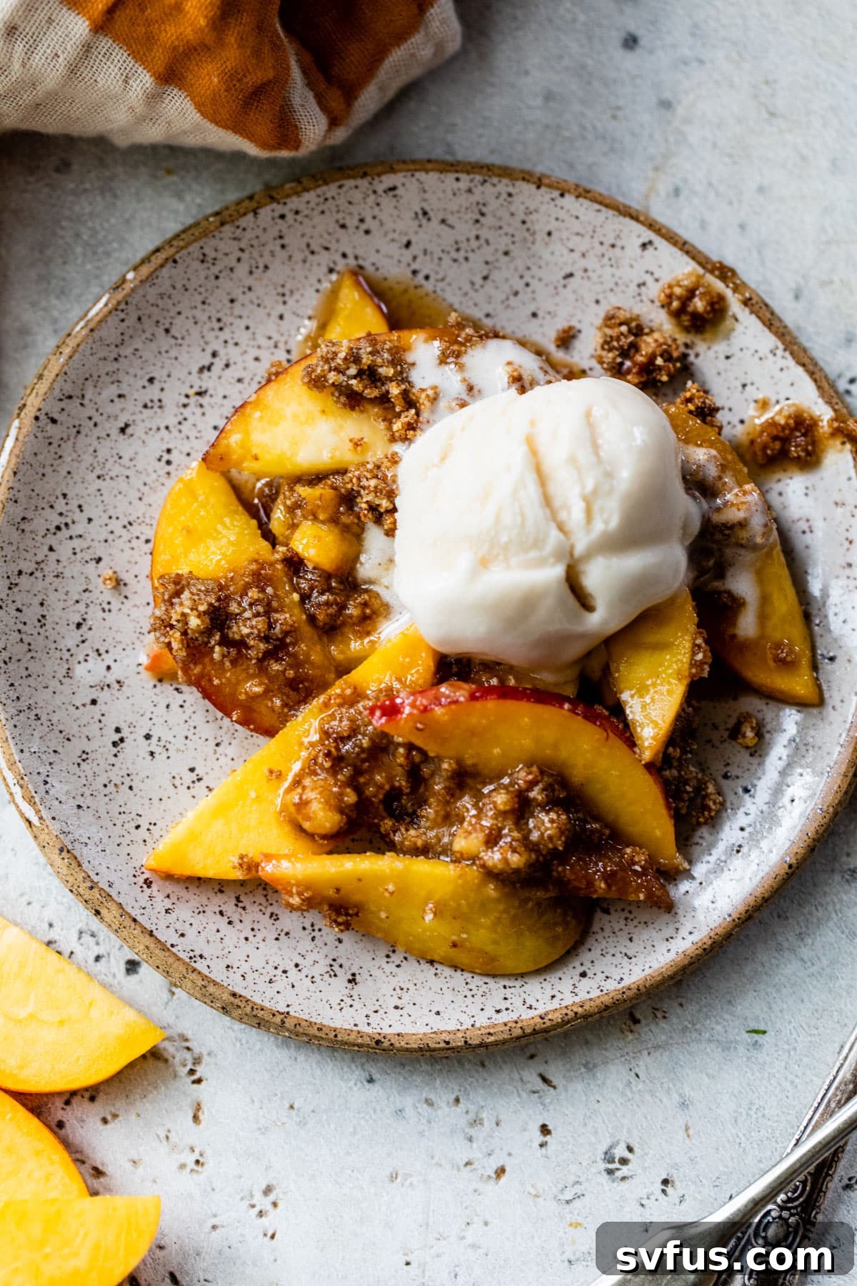 A serving of warm baked peach crisp topped with melting vanilla ice cream and a dusting of powdered sugar on a white plate.