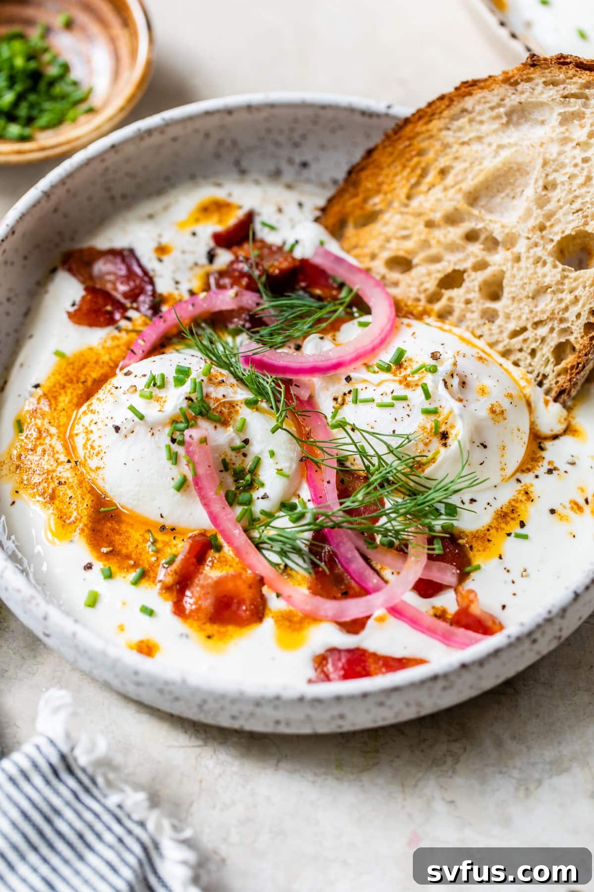 A beautifully arranged bowl of Turkish Eggs featuring a swirl of garlicky yogurt, perfectly poached eggs, crispy bacon, pickled red onions, and fresh herbs, served alongside toasted sourdough.