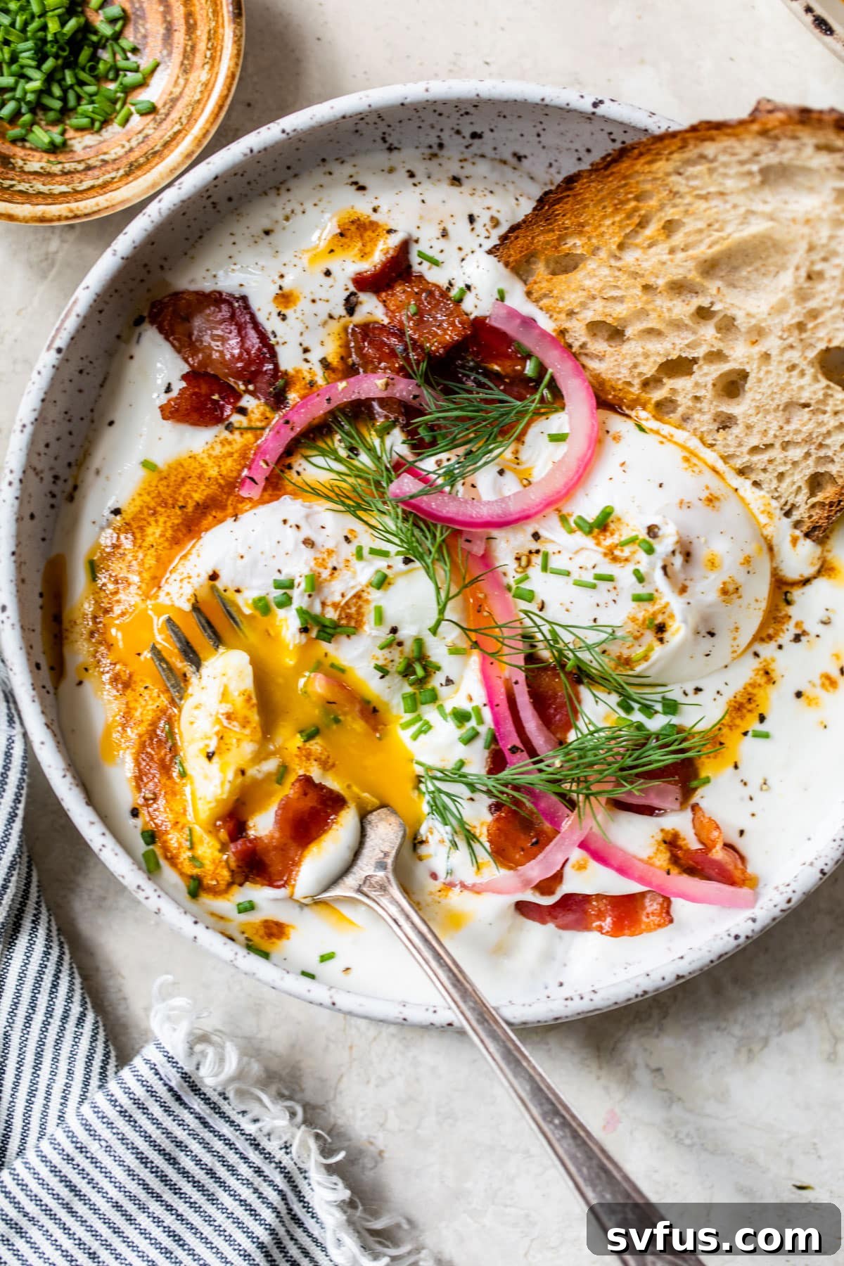 A large, inviting bowl of Turkish Eggs, generously topped with two poached eggs, crispy bacon, and a vibrant scattering of fresh herbs, showcasing the rich texture of the dish.