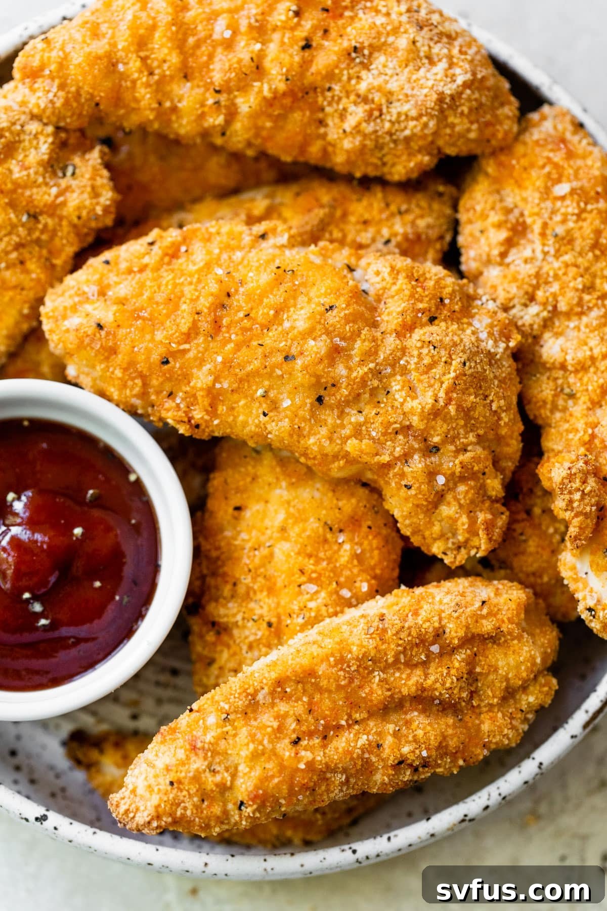 Close up of perfectly breaded and golden Whole30 chicken tenders in a serving bowl, with a small bowl of sugar-free ketchup on the side, ready for dipping.