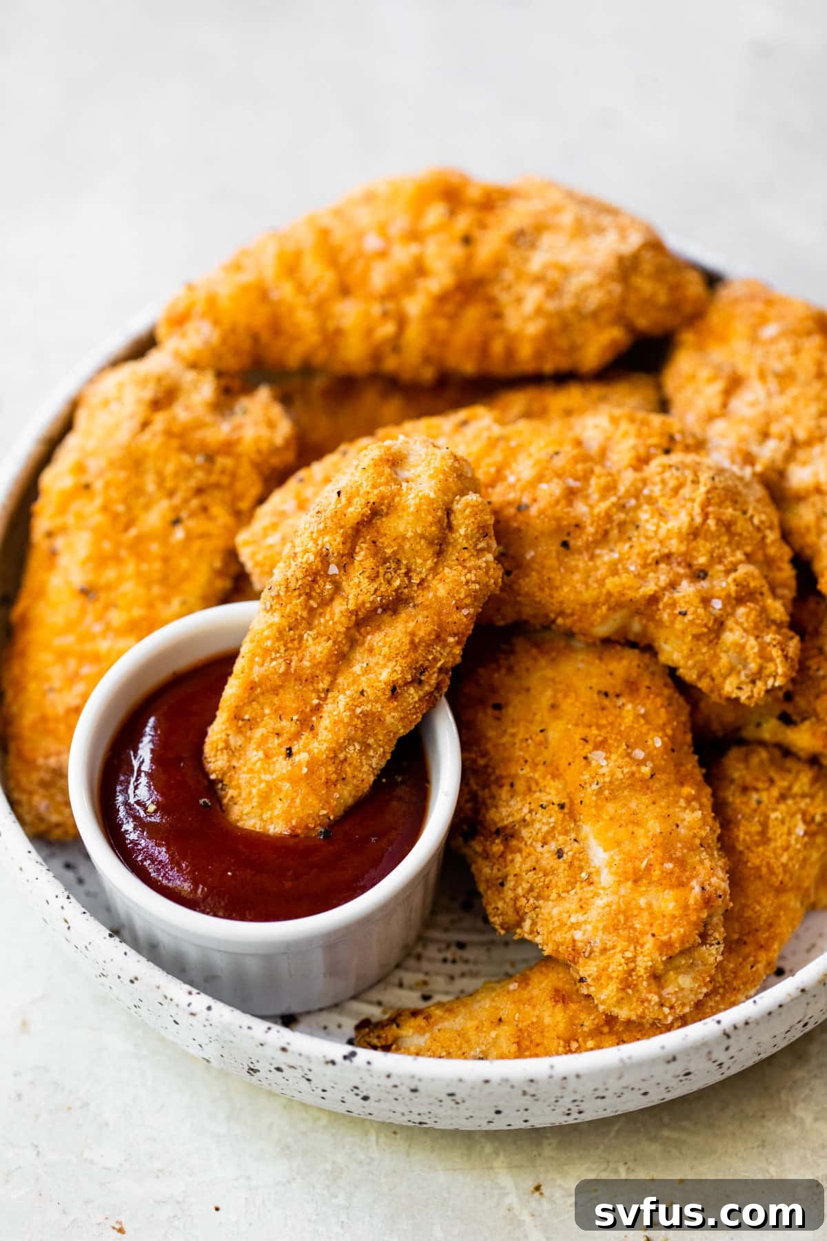 A bowl filled with golden-brown chicken tenders, with one tender being dipped into a small bowl of vibrant red ketchup, emphasizing the delicious pairing.