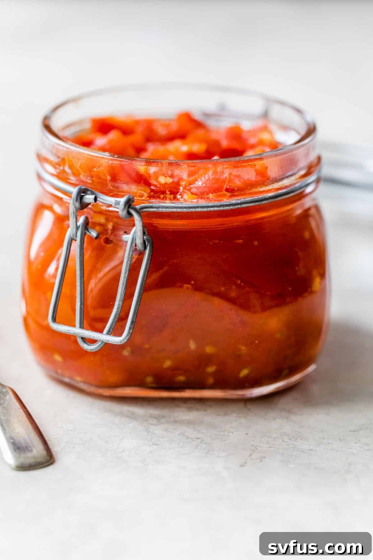 A short, clear glass jar filled with vibrant red homemade tomato relish, showcasing its chunky texture.