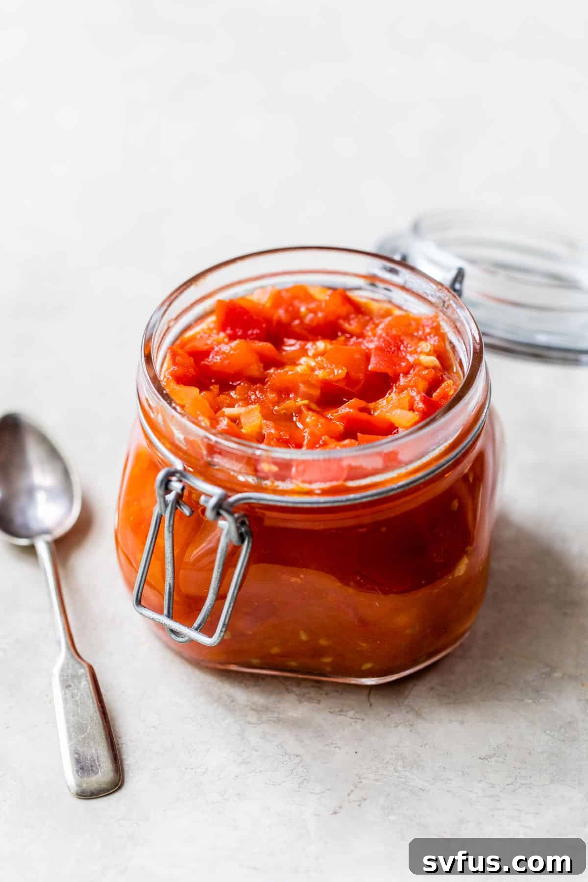 A glass jar filled with cooked diced tomatoes and herbs, next to a small wooden spoon, on a rustic wooden surface.