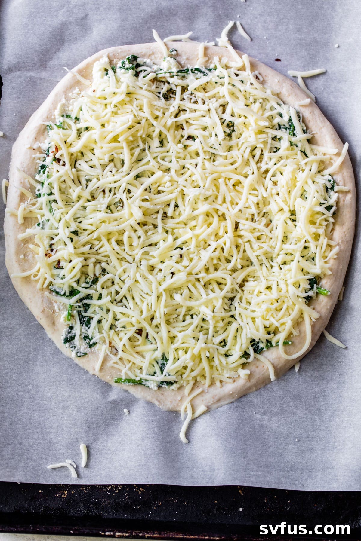 Unbaked pizza dough on parchment paper, generously topped with shredded mozzarella cheese, ready for the oven.