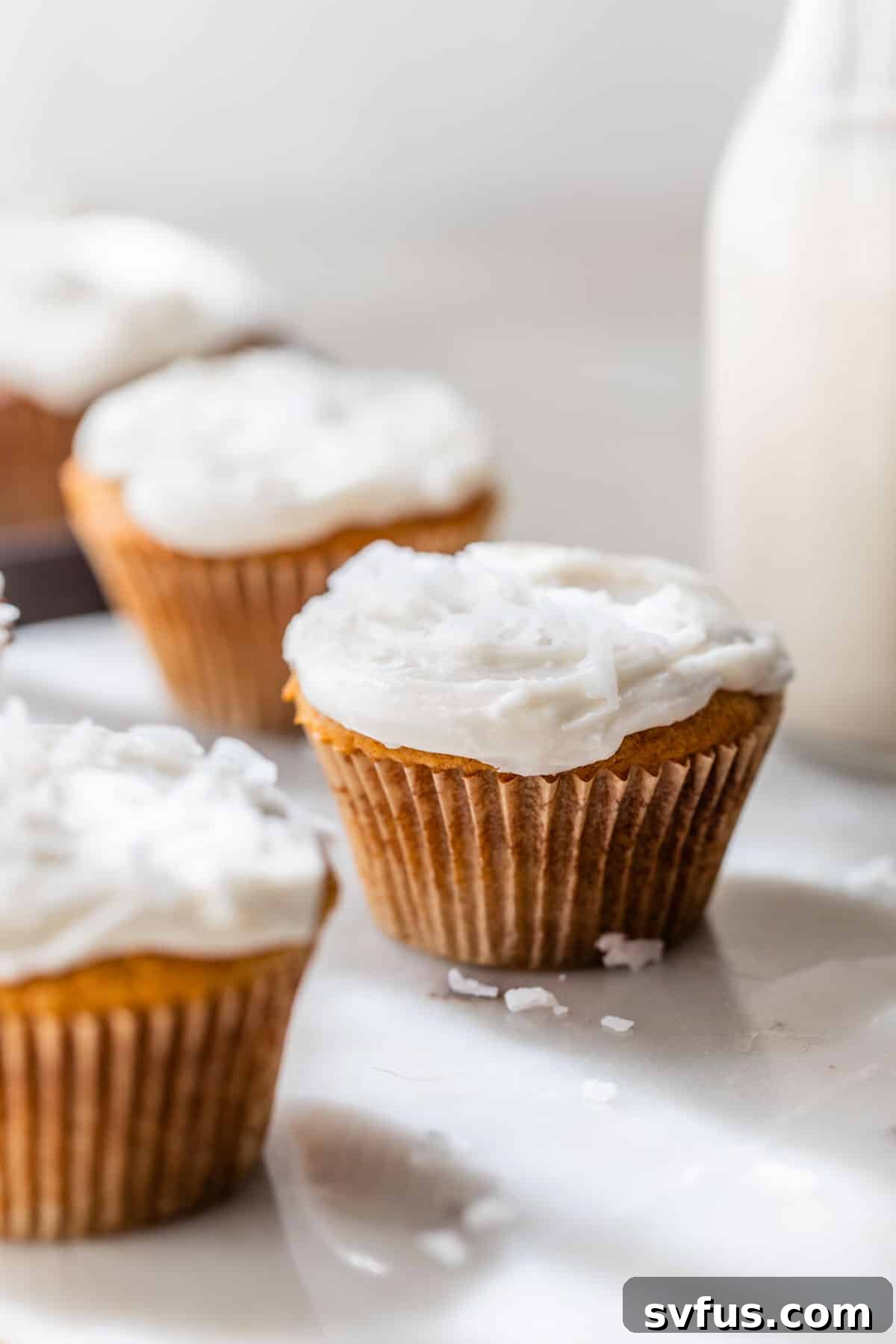cupcakes with white icing and shredded coconut on a table beside a glass of milk