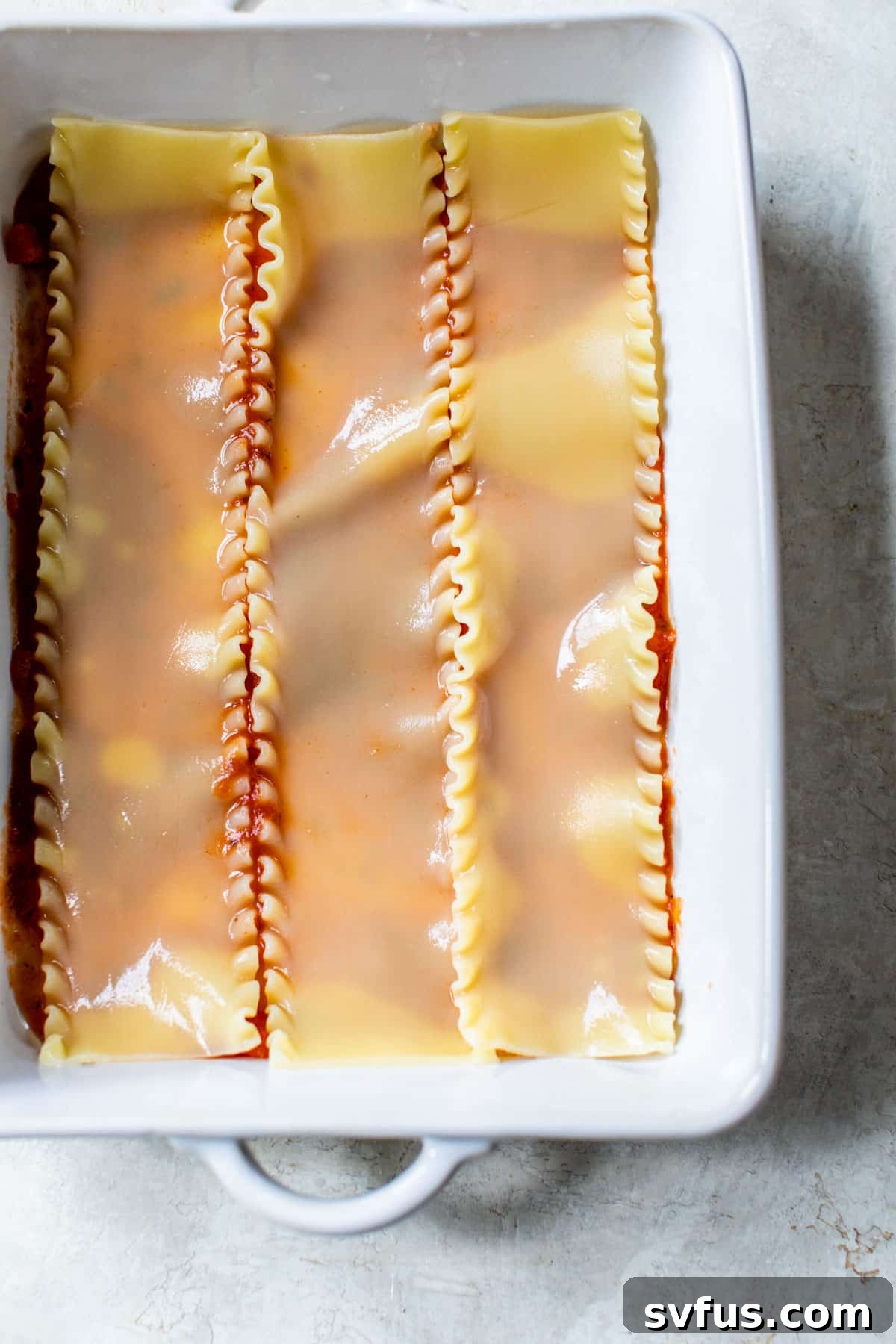 A rectangular baking dish with a base layer of vibrant red marinara sauce, topped with three perfectly placed lasagna noodles, ready for the next layer of delicious filling.