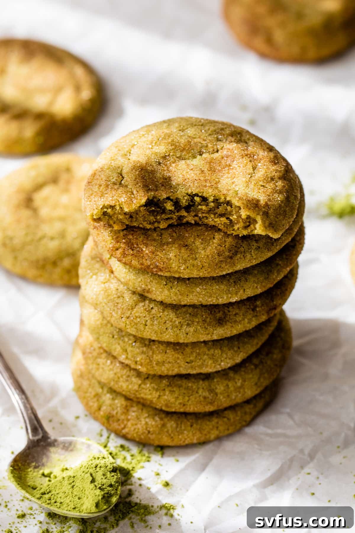 A stack of vibrant green matcha snickerdoodle cookies on parchment paper, next to a spoon filled with matcha powder.