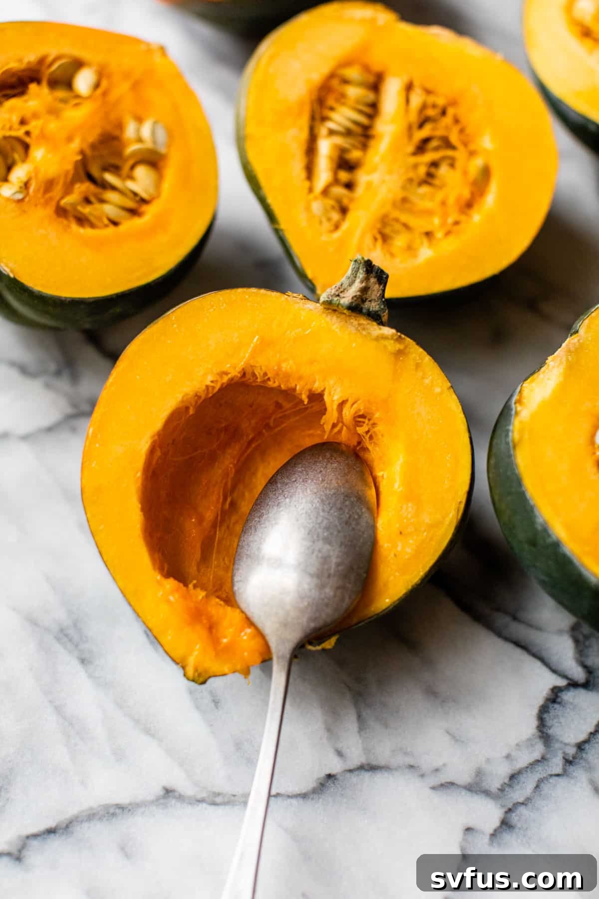 A spoon scooping out seeds and fibrous strands from the center of an acorn squash half, preparing it for stuffing.