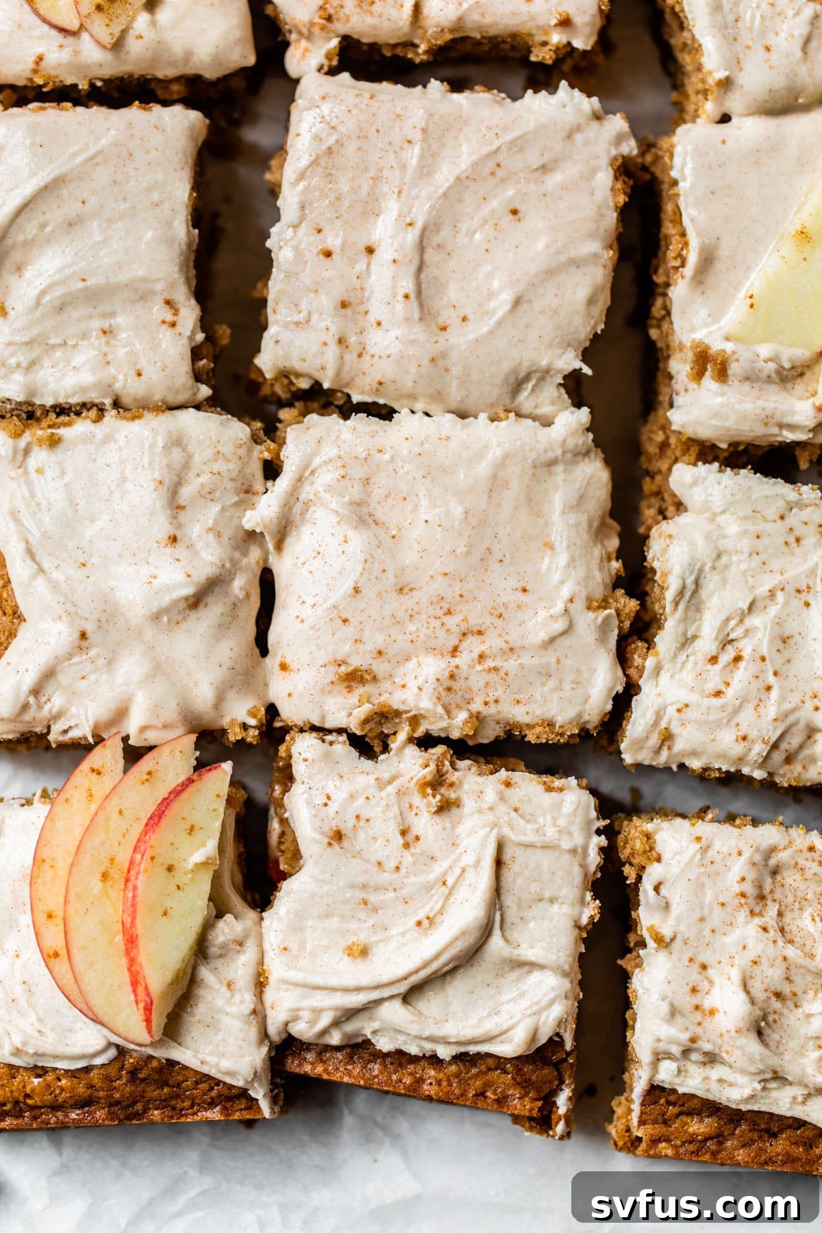 Square slices of Vegan Apple Cake with creamy white icing, topped with delicate apple slices and a sprinkle of cinnamon, arranged invitingly on a platter.