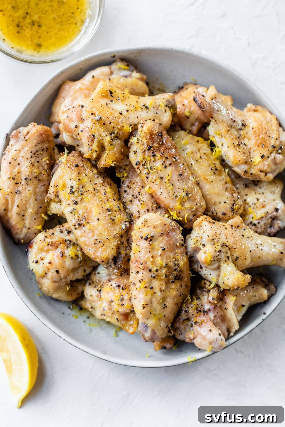 A bowl of glossy, sauced chicken wings generously topped with fresh lemon zest and cracked black pepper, next to a small bowl of melted butter.