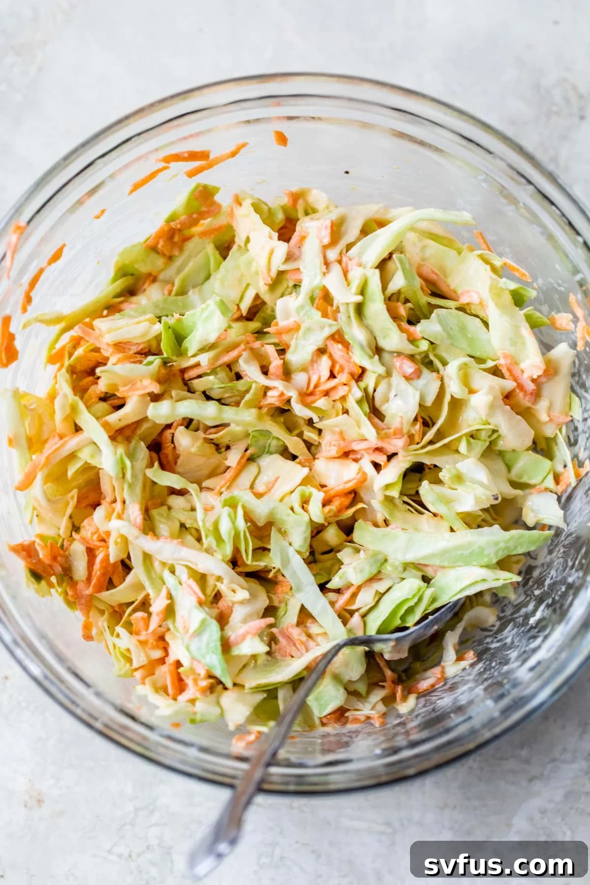 A large glass bowl filled with freshly prepared coleslaw, featuring shredded green cabbage and carrots mixed with a creamy dressing.