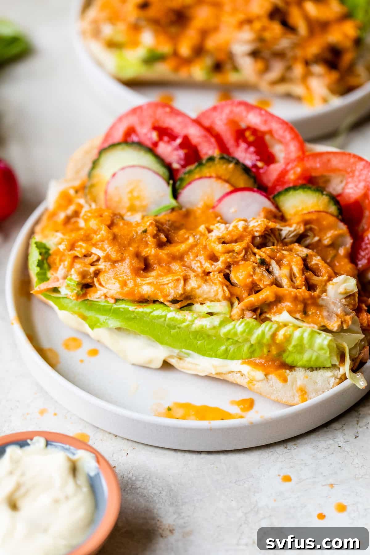 A close-up of a finished Panes con Pollo sandwich, showcasing layers of pulled chicken, crisp lettuce, sliced tomatoes, cucumbers, radishes, and a generous drizzle of the rich orange salsa.