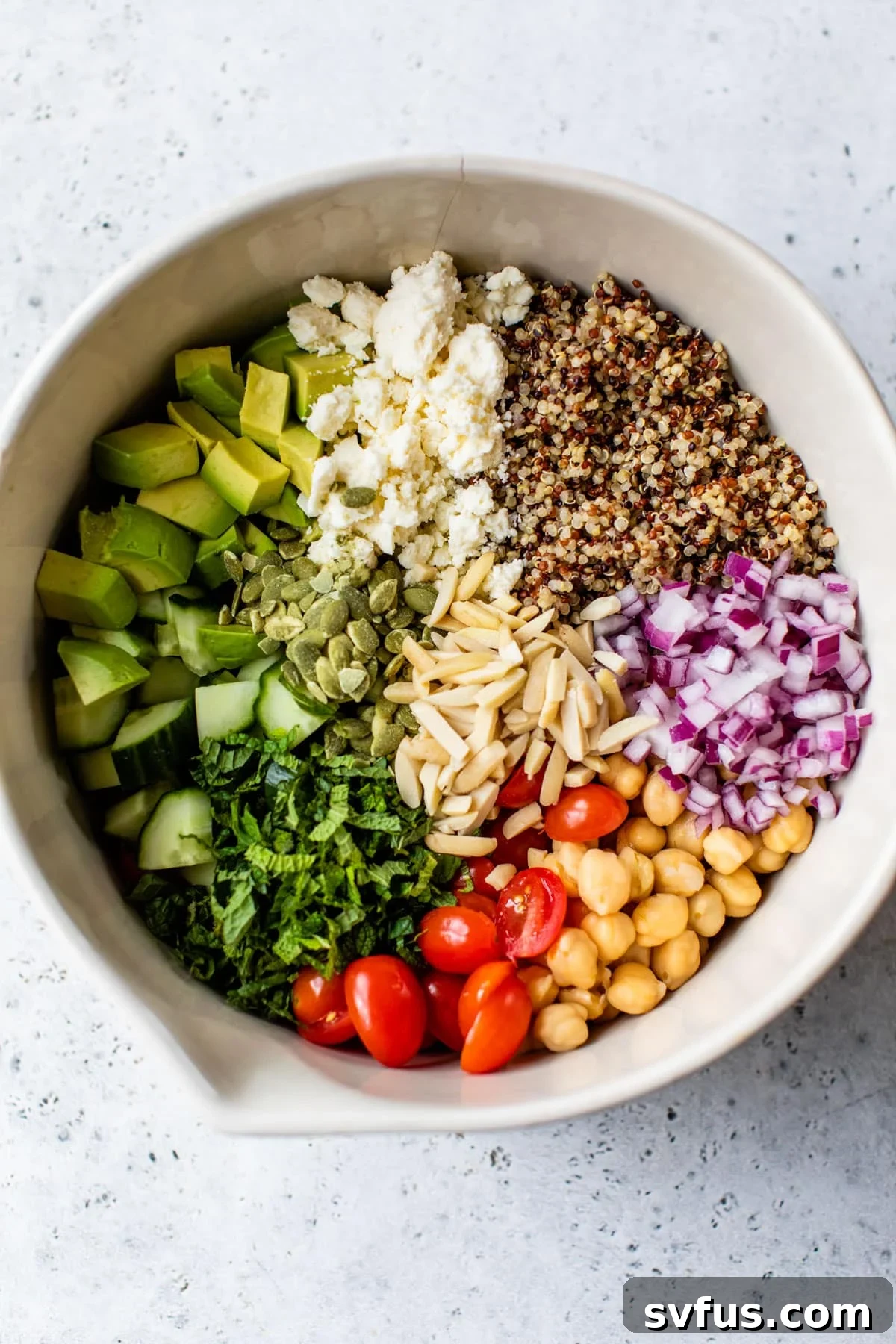 A large mixing bowl containing fluffy quinoa, colorful grape tomatoes, crisp cucumbers, diced red onion, chickpeas, and crumbled feta cheese, ready for the dressing.