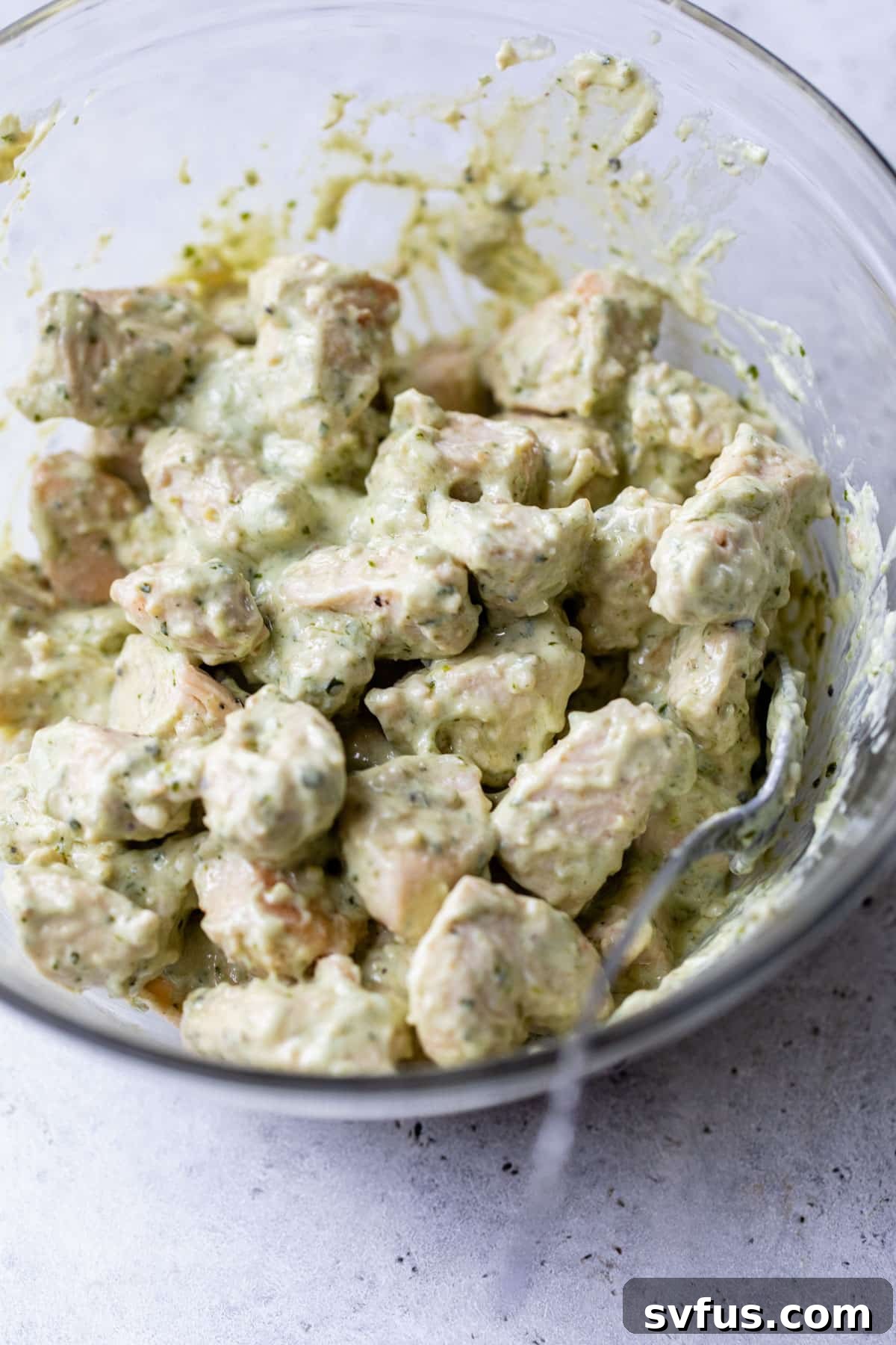 Shredded chicken pieces fully coated in vibrant green pesto dressing in a mixing bowl.