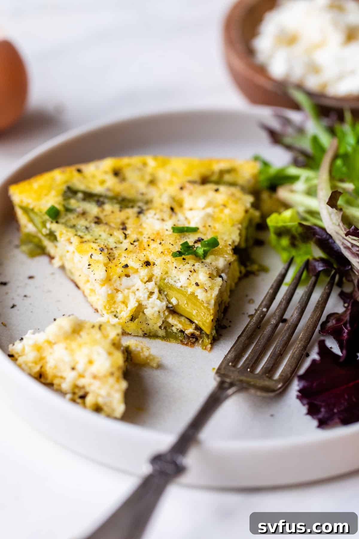 A slice of asparagus frittata on a white plate, with a fork cutting into a piece, highlighting its tender texture and vibrant green asparagus. The frittata features visible pieces of feta and a golden-brown top.