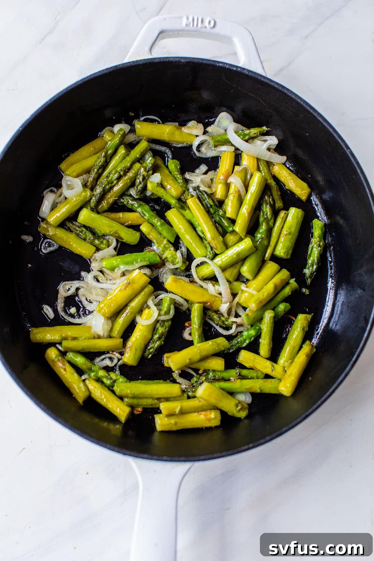 Sliced shallots and chopped asparagus are gently sautéing in a hot cast iron skillet, showcasing the cooking process before the egg mixture is added. The vegetables are vibrant and tender.