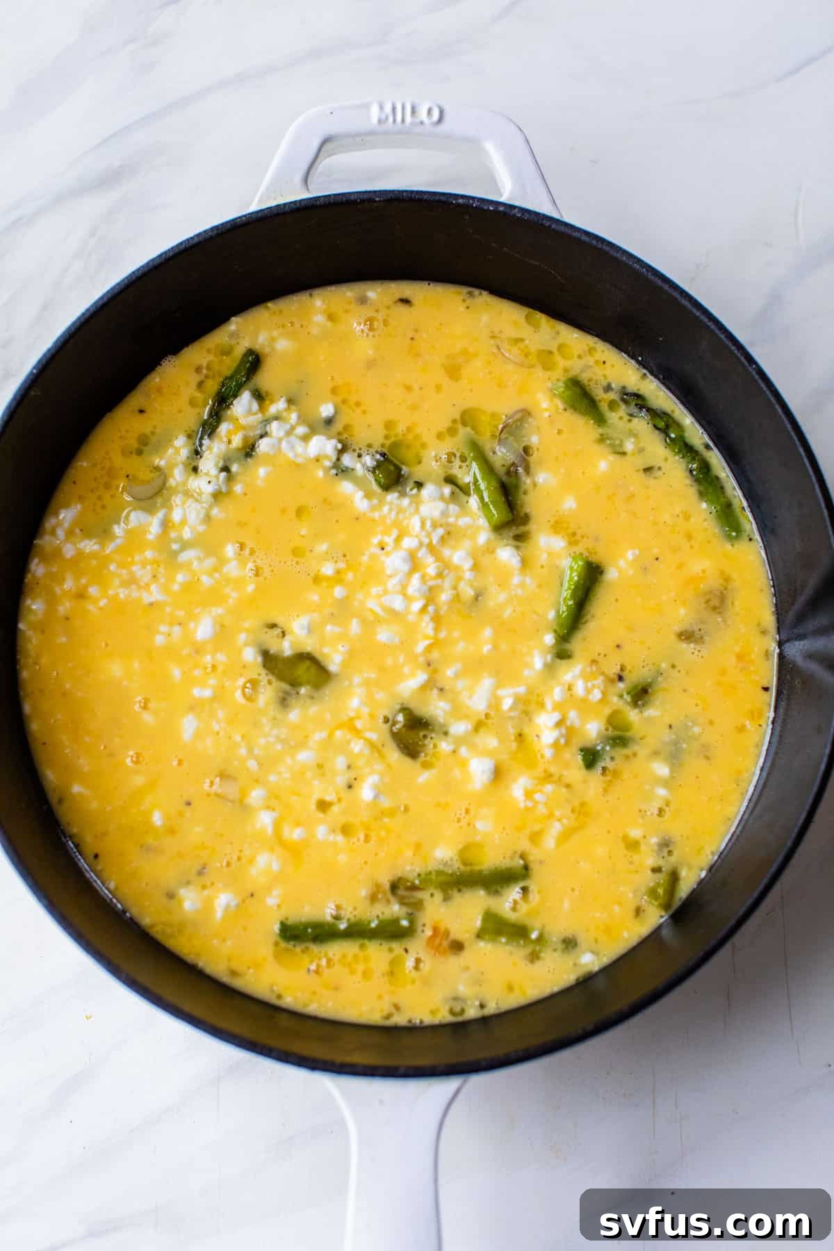 The vibrant asparagus and egg mixture, topped with crumbled feta cheese, gently cooking in an oven-safe skillet on the stovetop. The edges are just beginning to set, preparing it for baking.