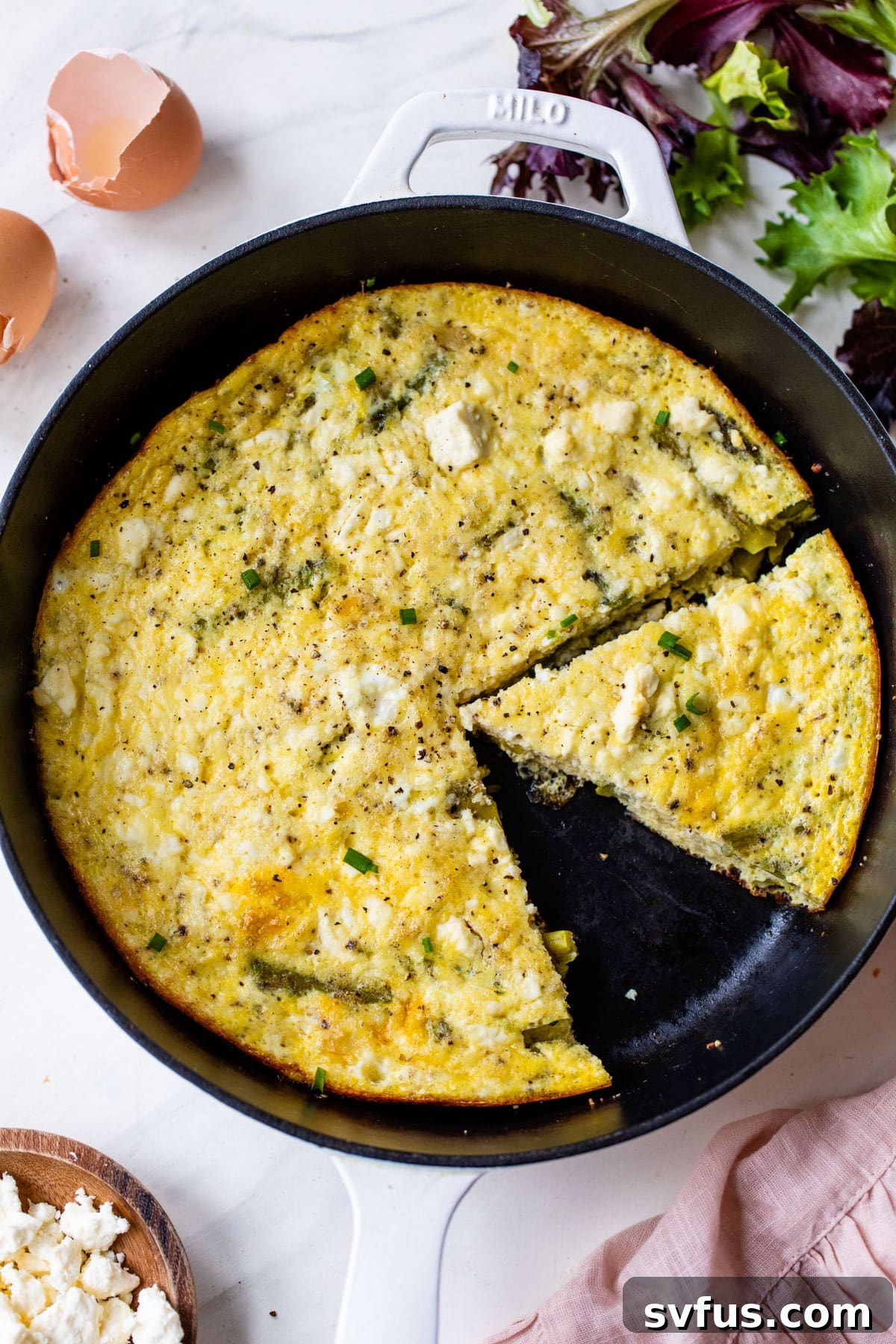 A freshly baked asparagus frittata in a cast iron skillet, with a slice already removed, showcasing its fluffy interior and golden-brown crust. The remaining wedges are ready to be served.