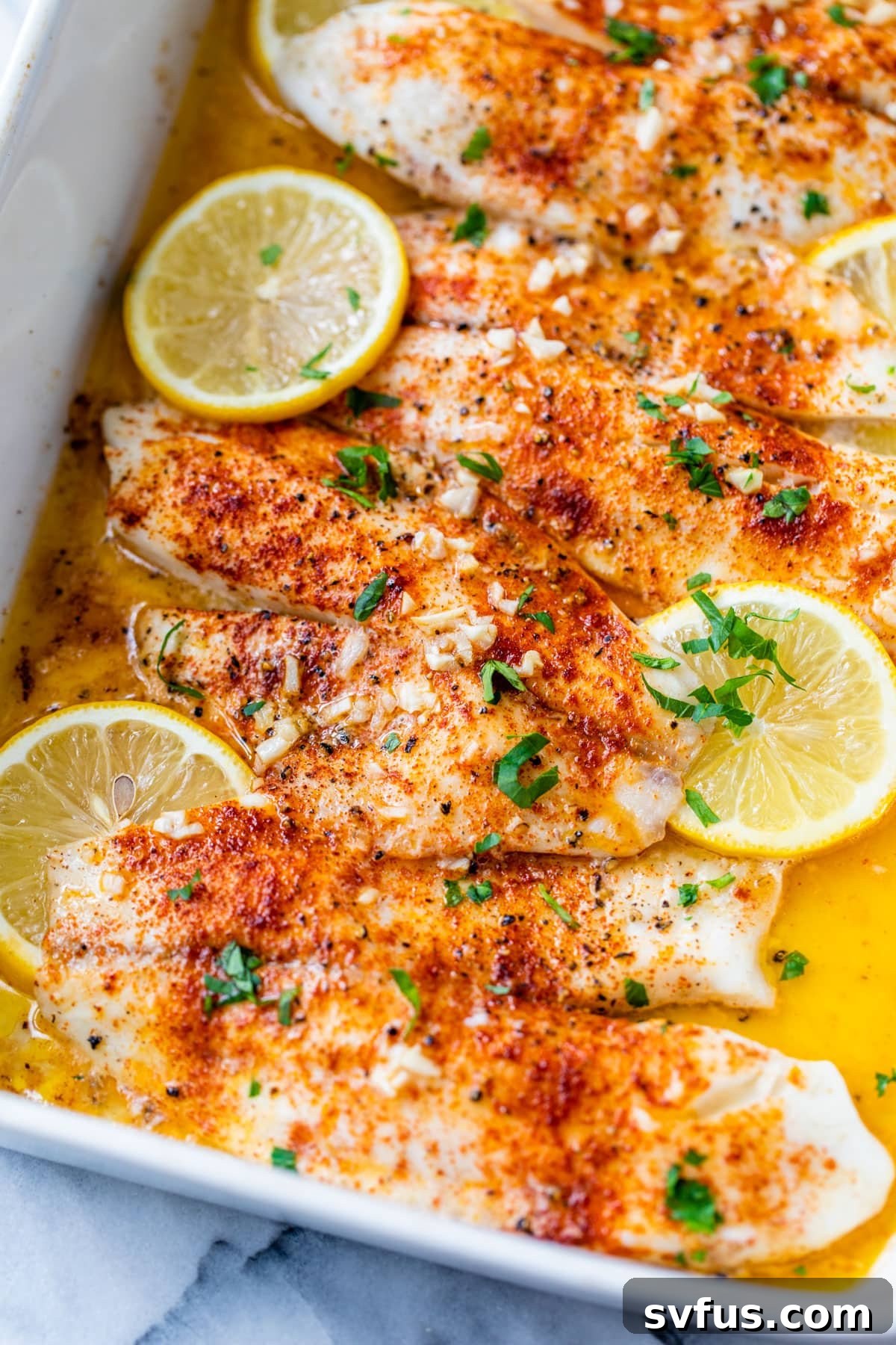 Baked tilapia fillets in a baking dish, garnished with fresh lemon slices and parsley
