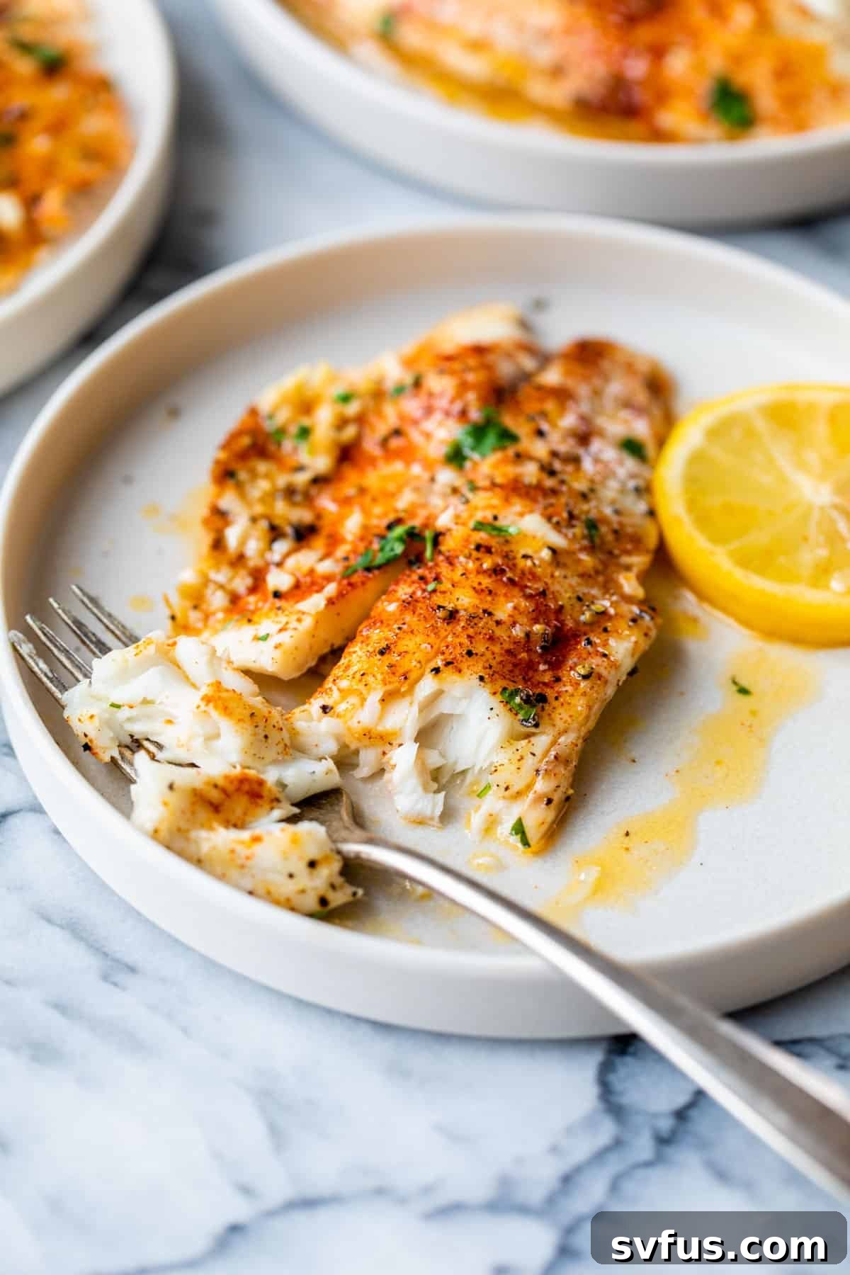 A close-up of baked tilapia on a white plate with a fork, showing its flaky texture and lemon garnish