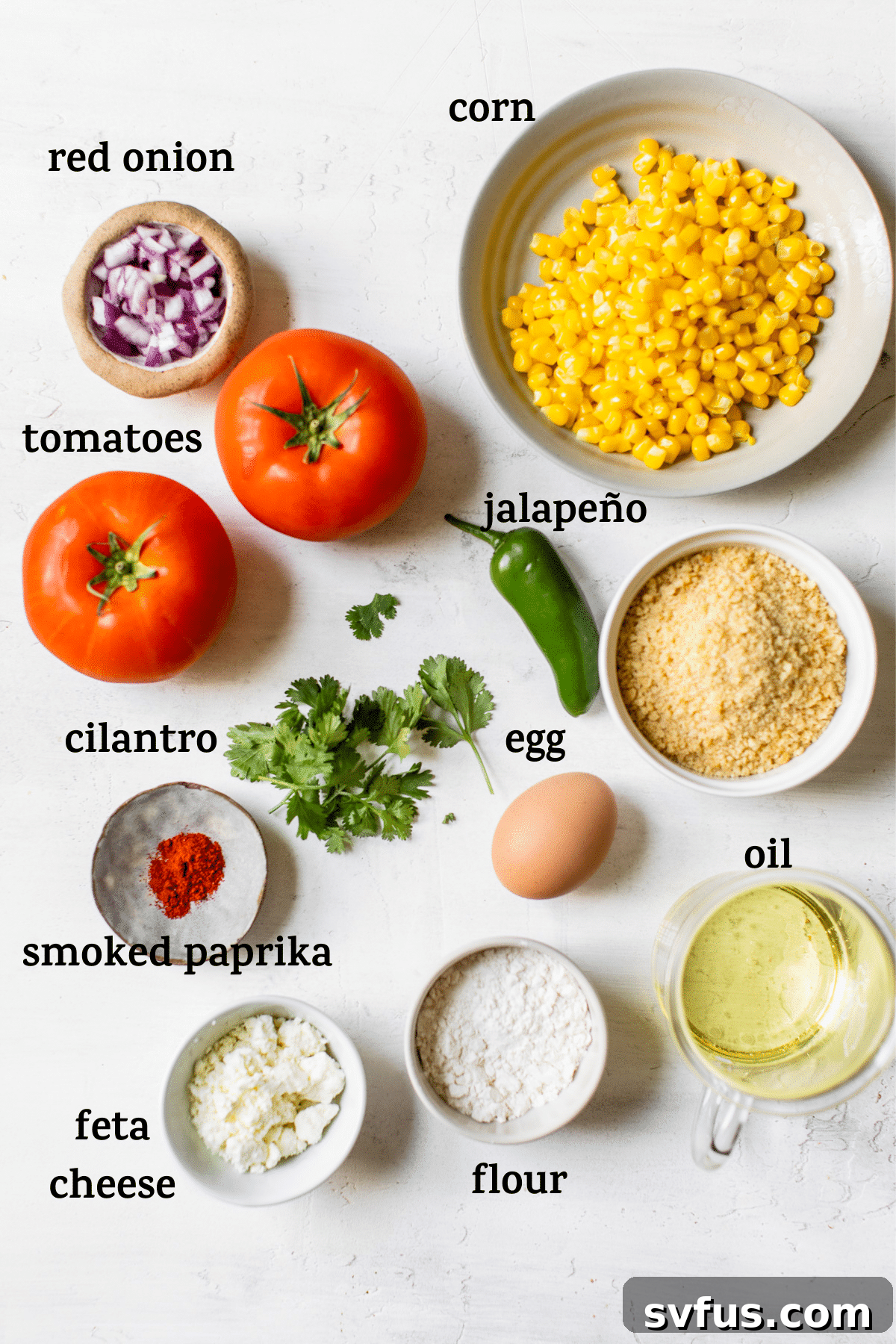 A collection of fresh ingredients on a wooden table, including beefsteak tomatoes, eggs, Panko breadcrumbs, olive oil, corn, feta cheese, jalapeño, red onion, and cilantro.