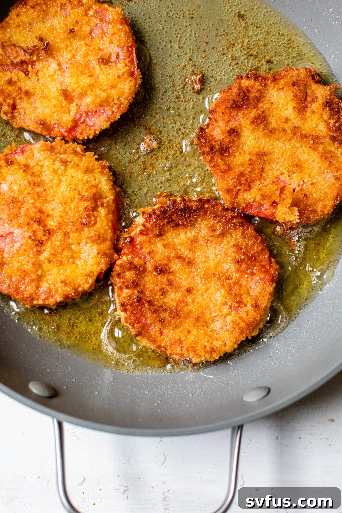 Several breaded tomato slices sizzling and turning golden brown in hot oil within a deep skillet.