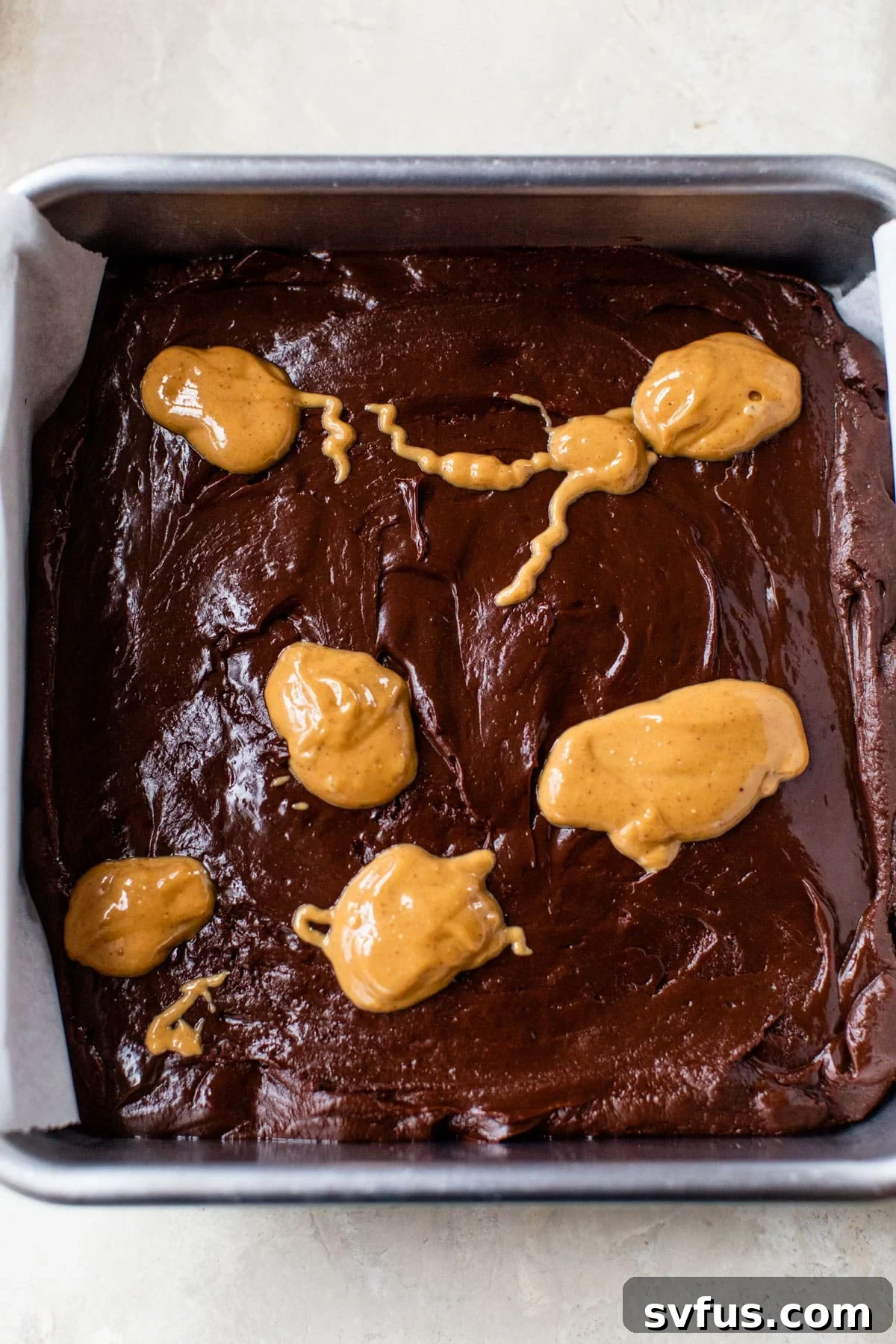 A square baking pan filled with smooth chocolate mixture, topped with dollops of creamy peanut butter ready for swirling.