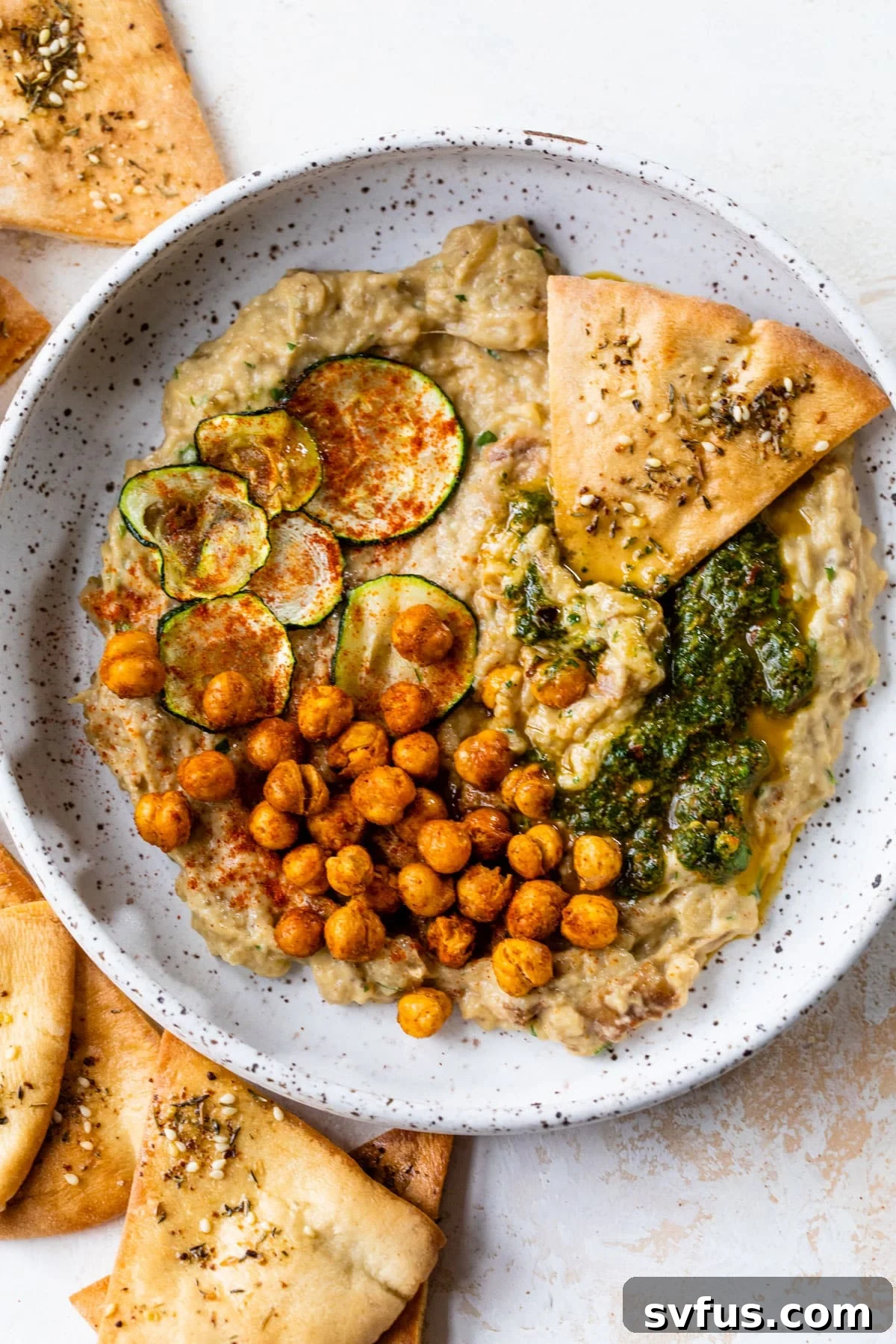 A beautifully presented bowl of creamy baba ganoush, generously topped with crispy roasted chickpeas, a vibrant green chermoula sauce, delicate slices of zucchini, and accompanied by warm pita chips.