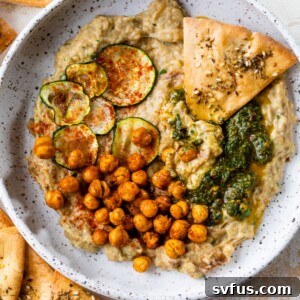 a bowl of baba ganoush topped with chickpeas, green sauce, sliced zucchini, and pita chips