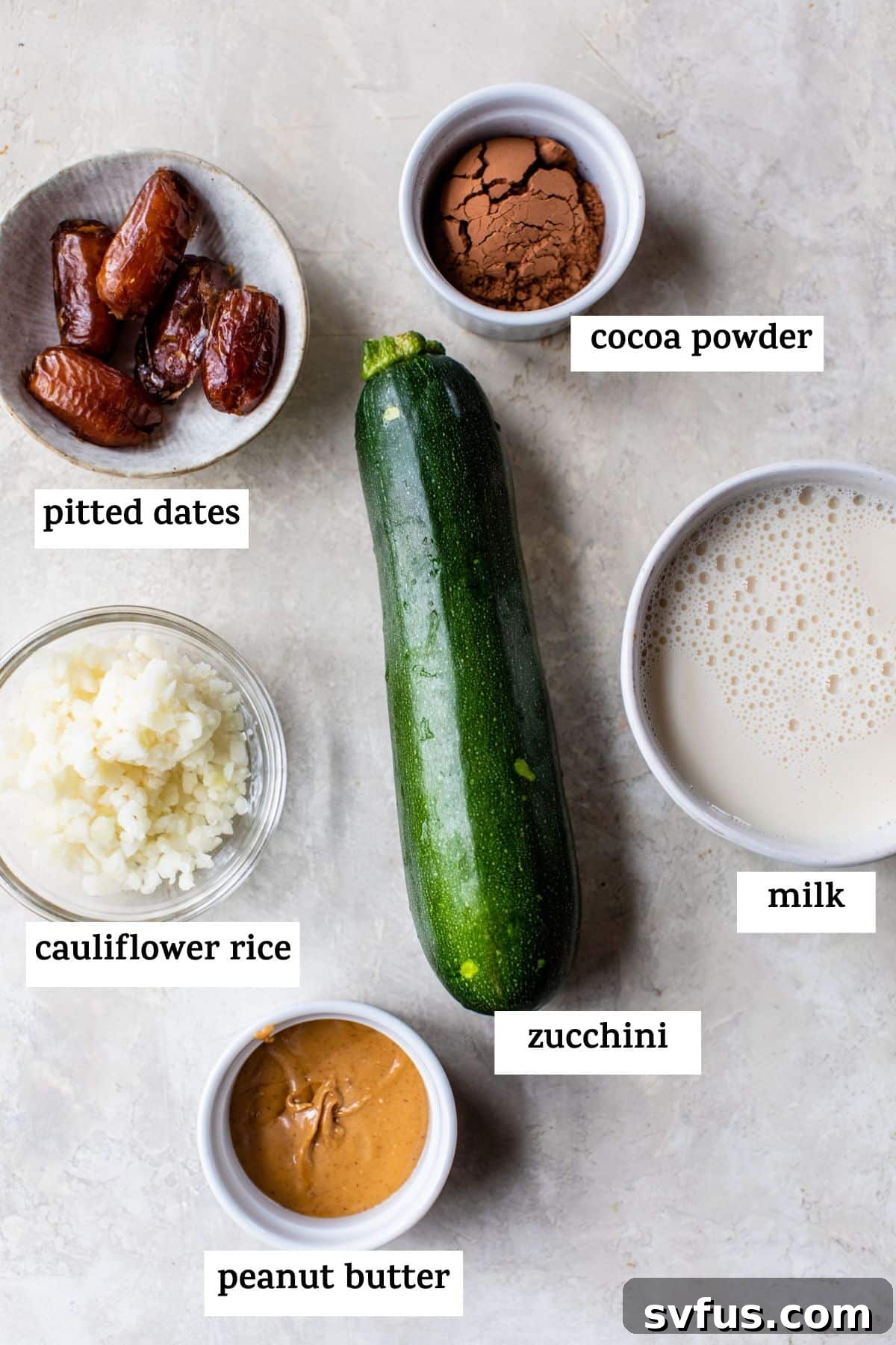 All six key ingredients for the healthy chocolate zucchini smoothie displayed: pitted dates, cauliflower rice, peanut butter, chopped zucchini, milk, and cacao powder