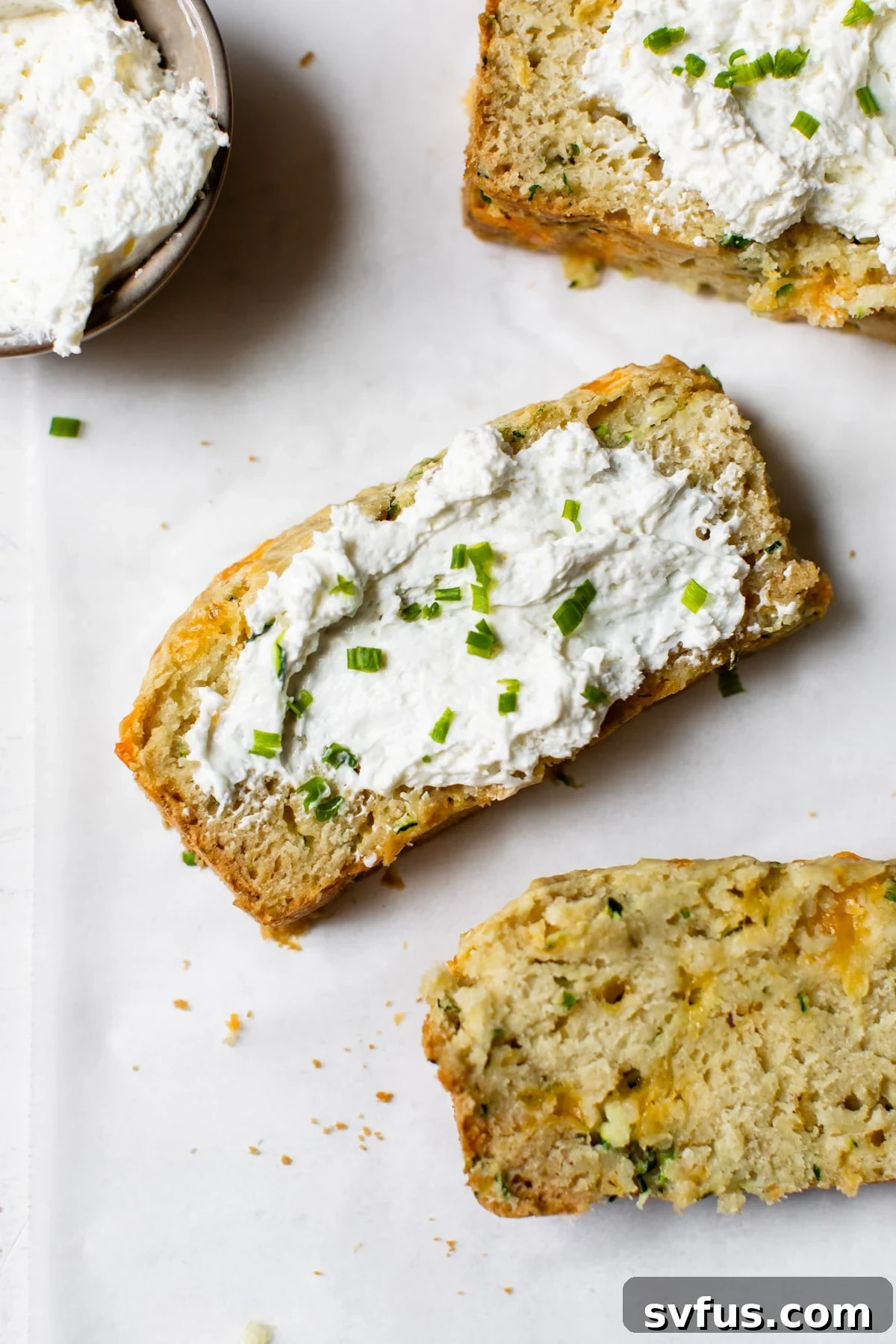 Multiple slices of golden brown savory zucchini bread, each topped with a generous dollop of creamy white chive cream cheese, arranged enticingly on a cooling rack