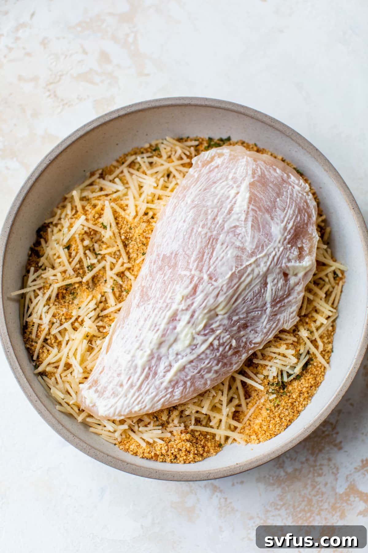 Raw chicken breasts, alongside separate bowls of mayonnaise and a mixture of breadcrumbs and parmesan cheese, ready for coating.